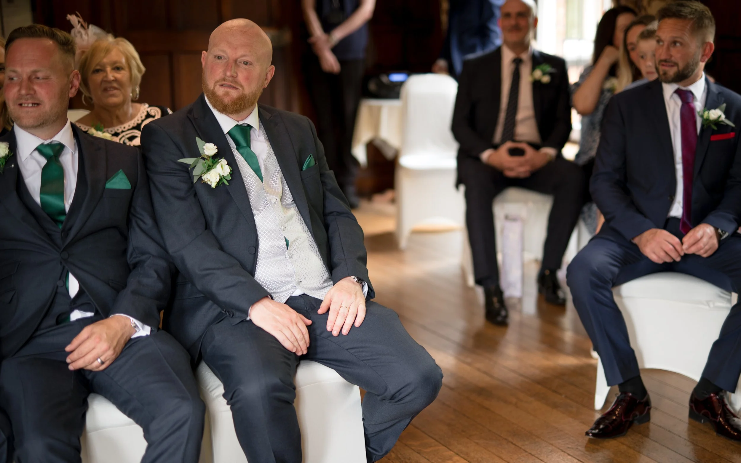 A group of people dressed in formal attire, sitting on white chairs in a wooden-floored room, attending a wedding or ceremony. The focus is on two men with boutonnières, one with a green tie and the other with a maroon tie, sitting in the front row.