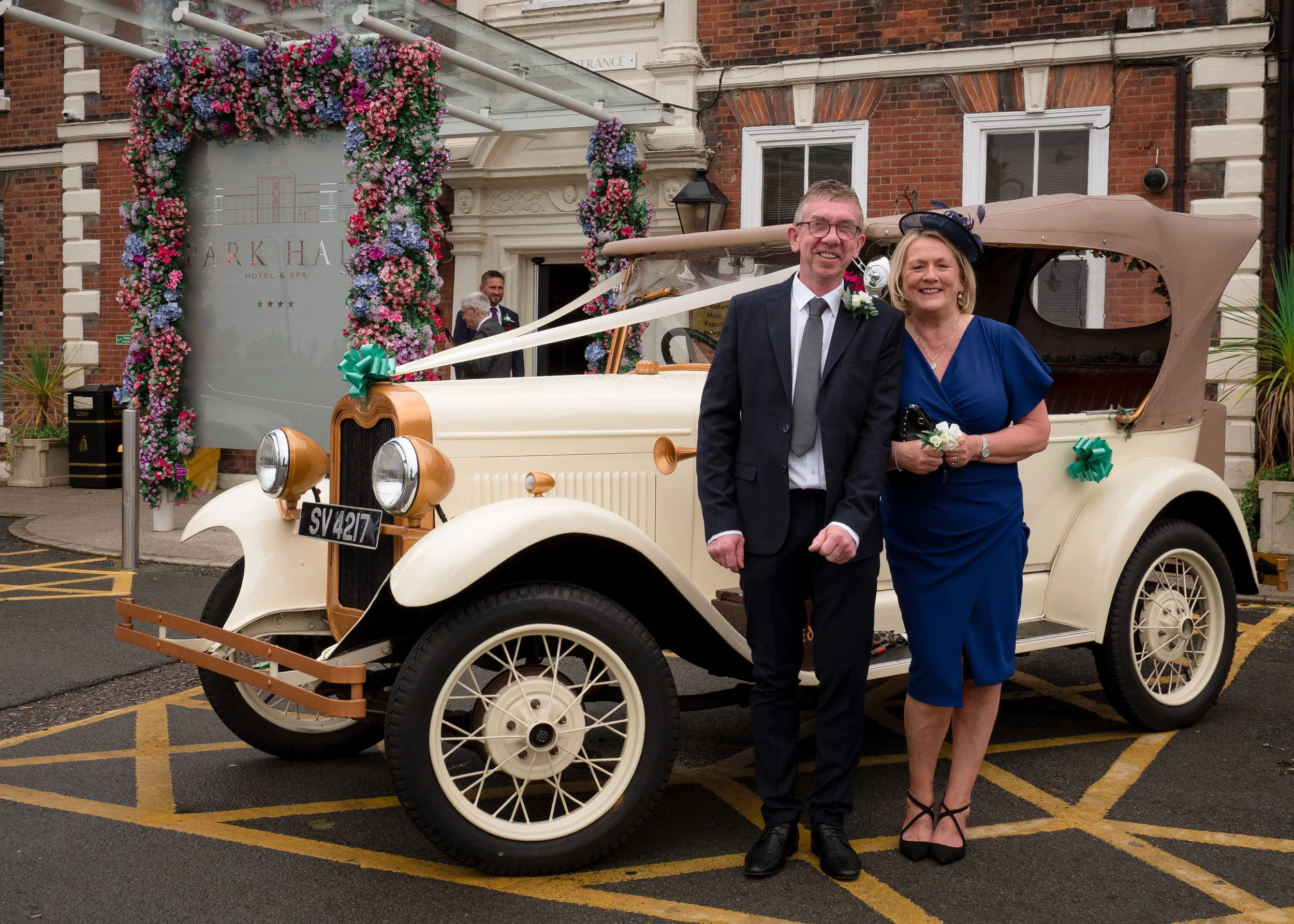 A happy man and woman stand in front of a vintage car decorated with ribbons and flowers outside a building entrance with a floral arch. The man is wearing glasses, a dark suit, and a boutonniere. The woman is smiling, dressed in a blue dress and a s