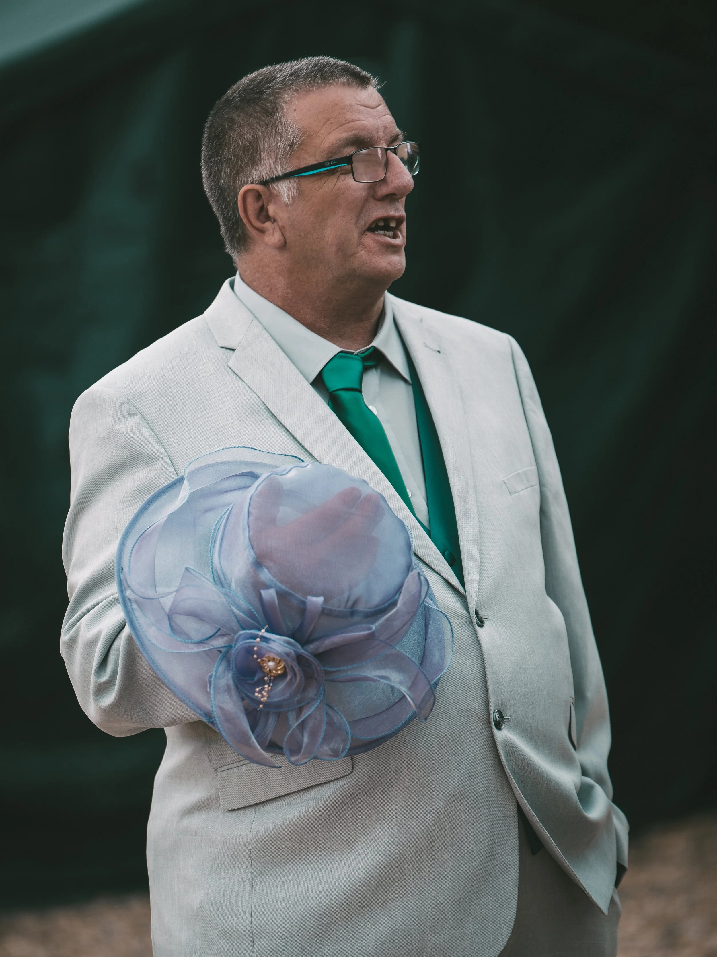 A man dressed in a white suit with a green tie, wearing glasses, holding a clear umbrella with a decorative attachment, and speaking outdoors.