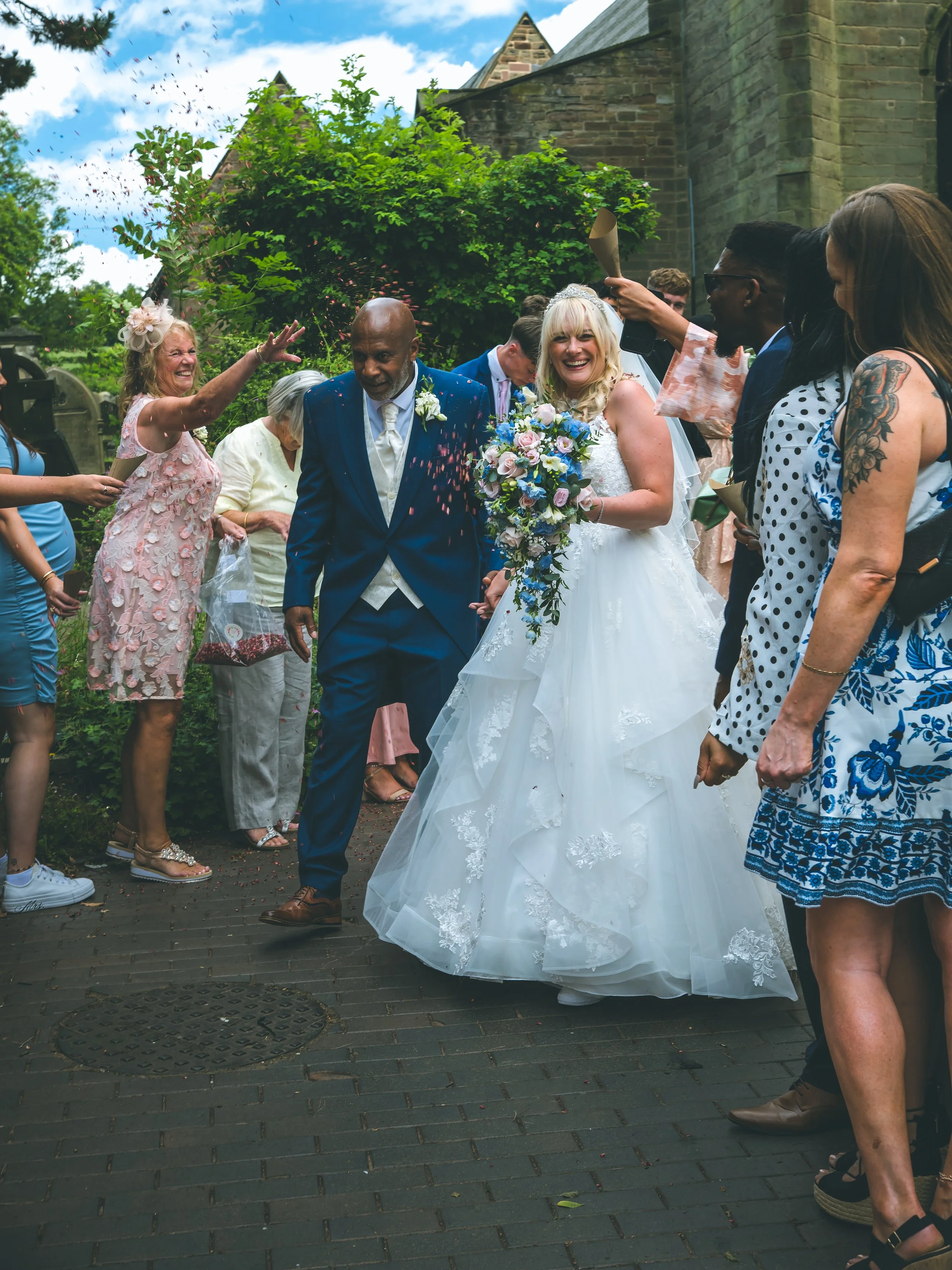 A bride in a white wedding dress holding a bouquet of flowers smiling while walking outdoors surrounded by guests, some throwing confetti, near a stone building under a blue sky.