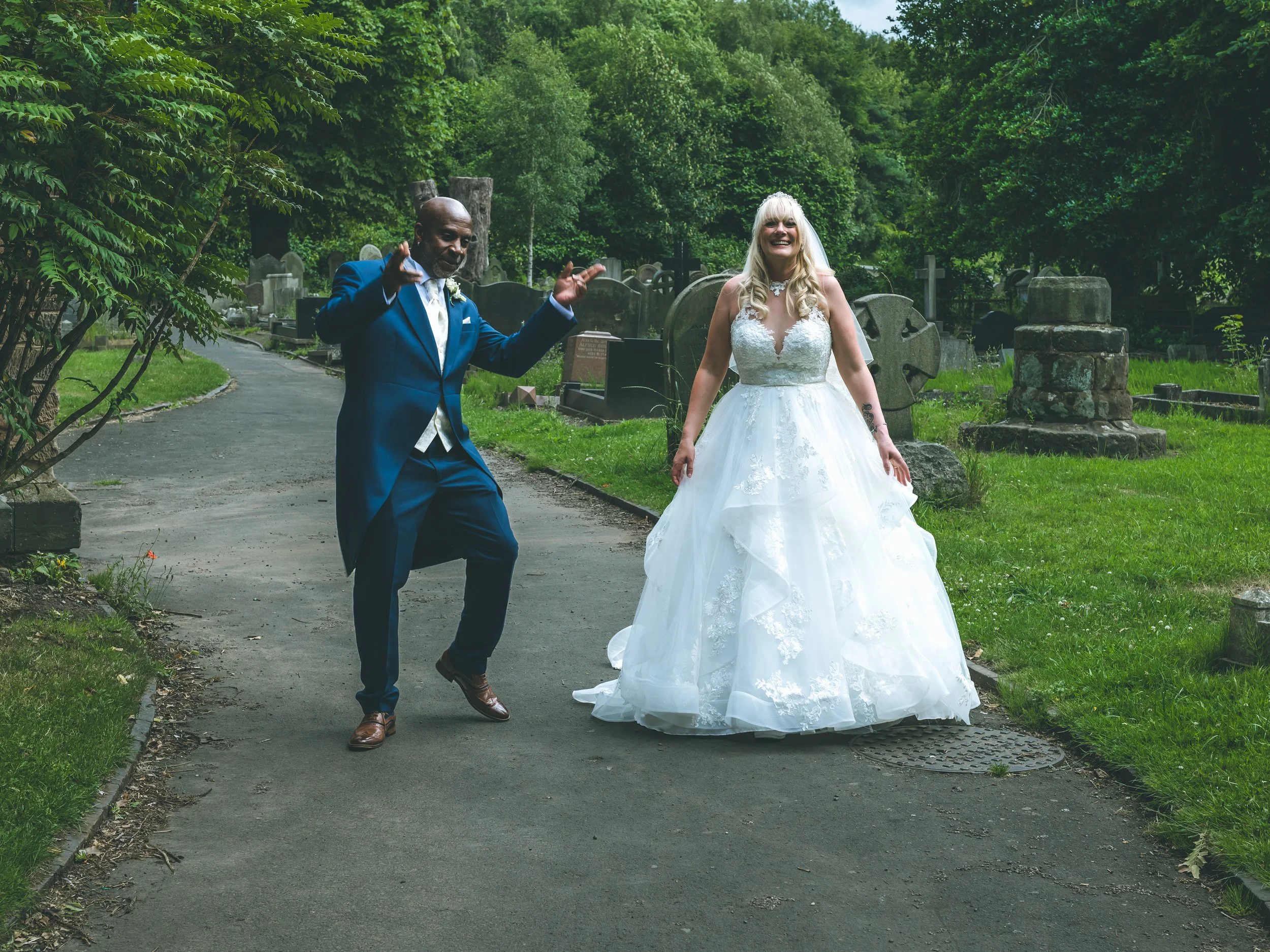 A bride in a white wedding dress walking in a cemetery with a man in a blue suit, surrounded by green trees and old tombstones.