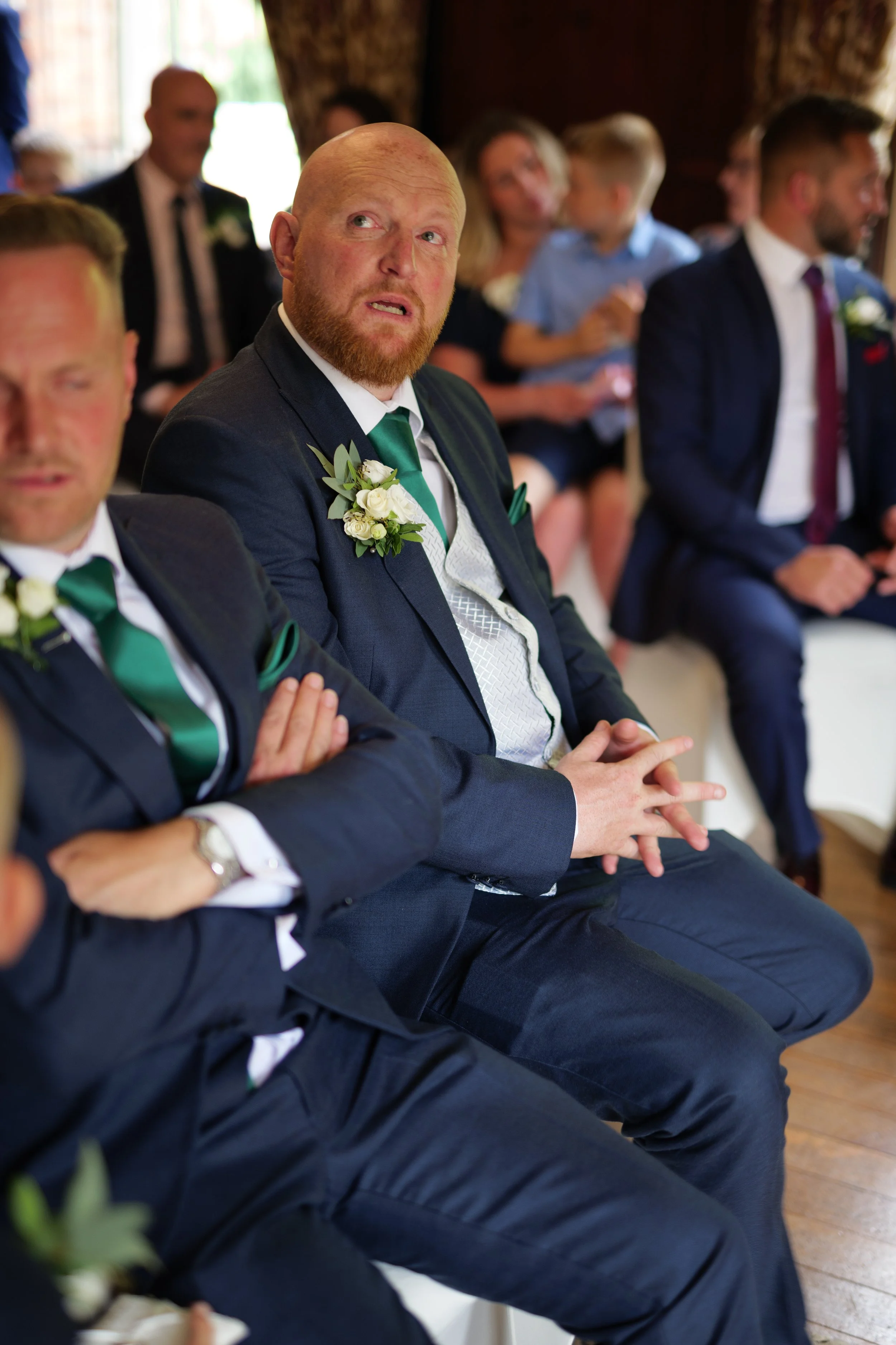 A group of people dressed in formal suits attending a wedding, with one man visibly emotional, wearing a dark suit, white shirt, green tie, and a flower boutonniere, seated in the front.