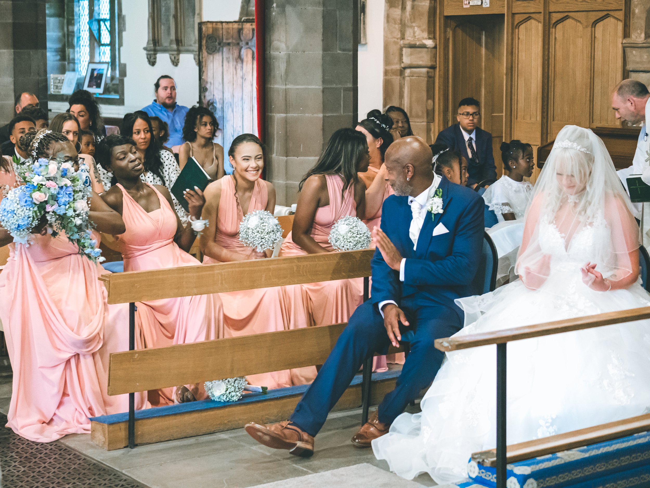A diverse group of wedding guests, including women in pink dresses and a bride in a white gown with a veil, seated inside a church during a wedding ceremony. The groom, dressed in a blue suit, is sitting and praying.