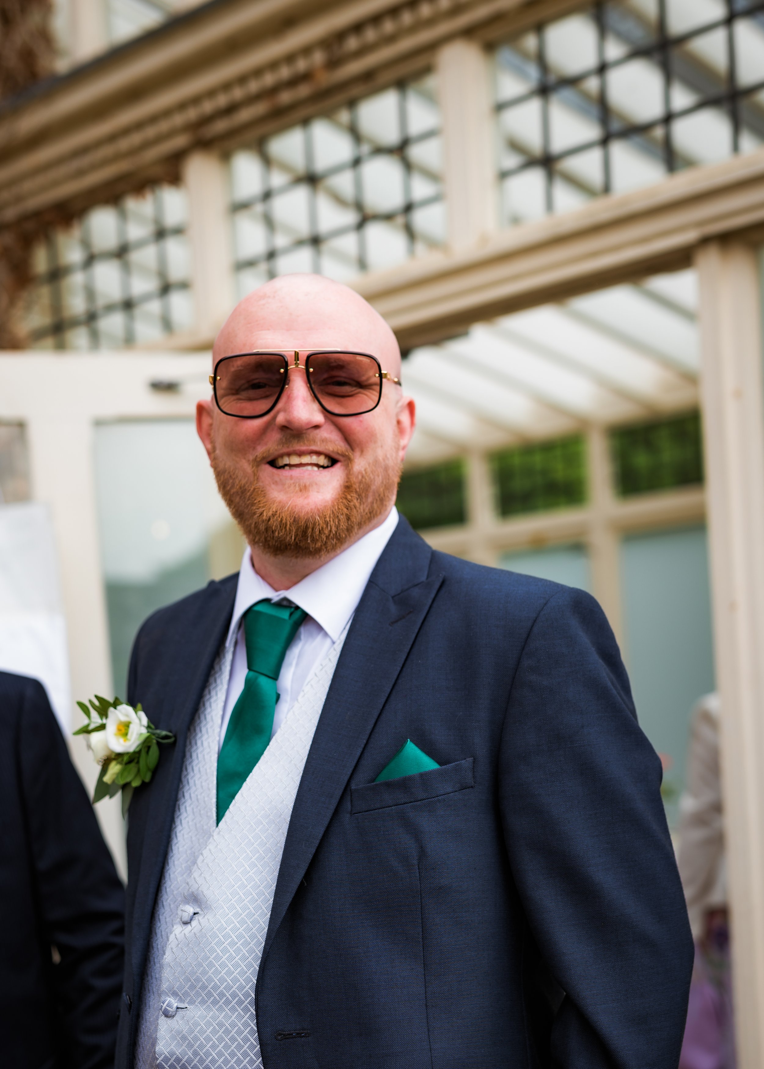 Close-up of a smiling man wearing a dark blue suit, white shirt, green tie, dark sunglasses, and a white boutonniere, at an outdoor event with a glass and wood structure in the background.