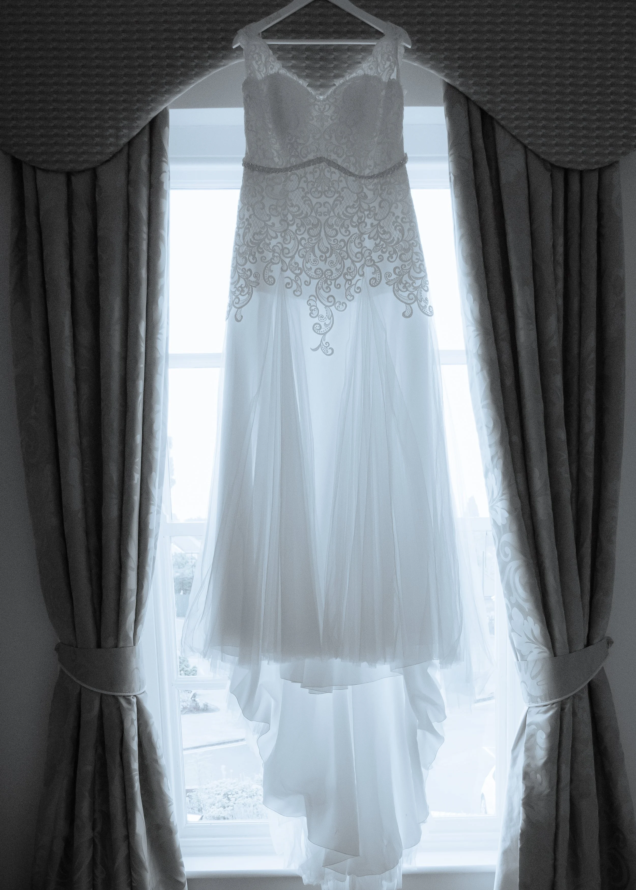 A wedding dress hanging from a hanger in front of a window with curtains, backlit by natural daylight.