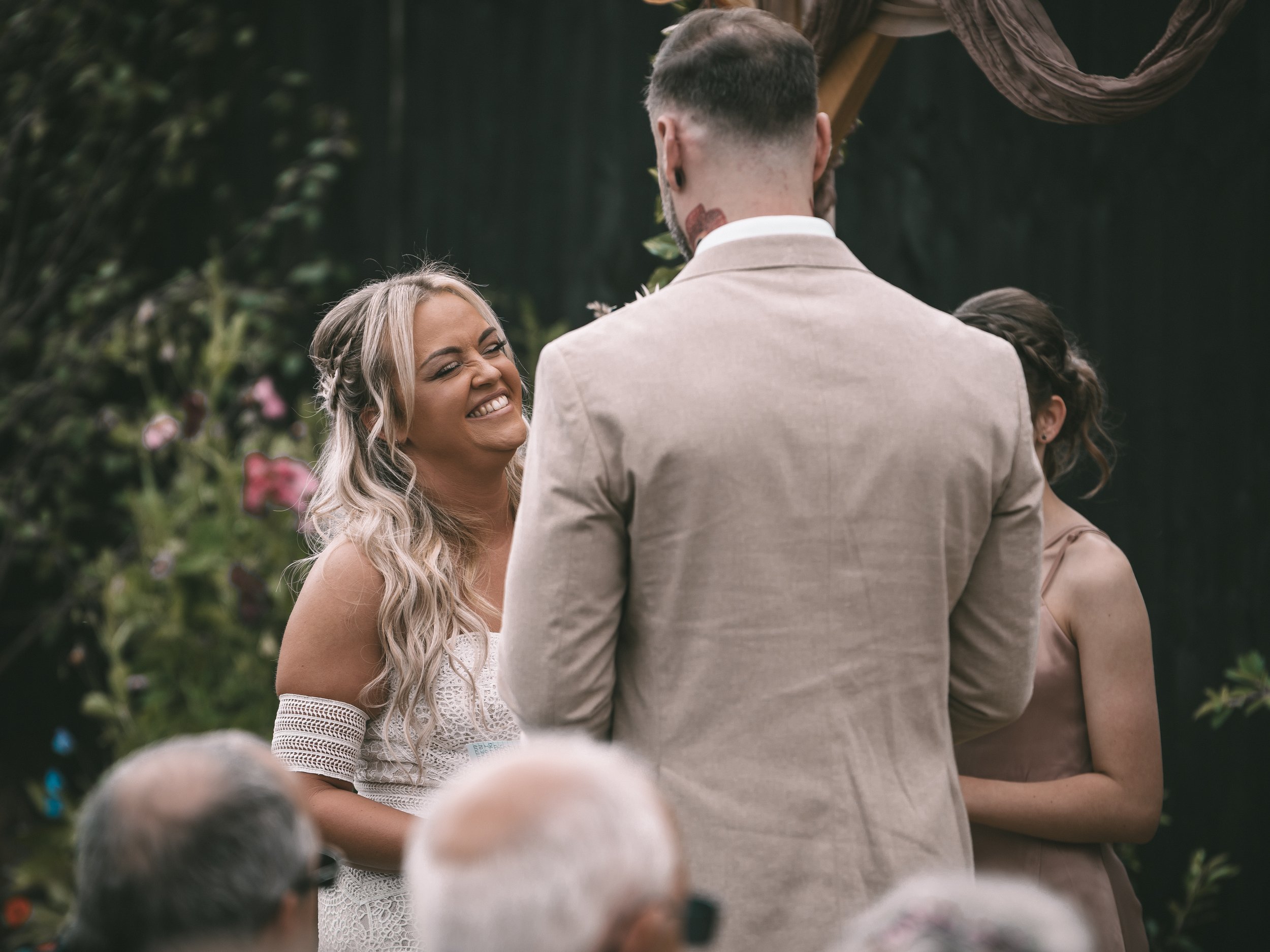 A woman with long blonde hair, wearing a white dress with lace details, smiling happily at a man during an outdoor wedding ceremony surrounded by greenery and flowers.