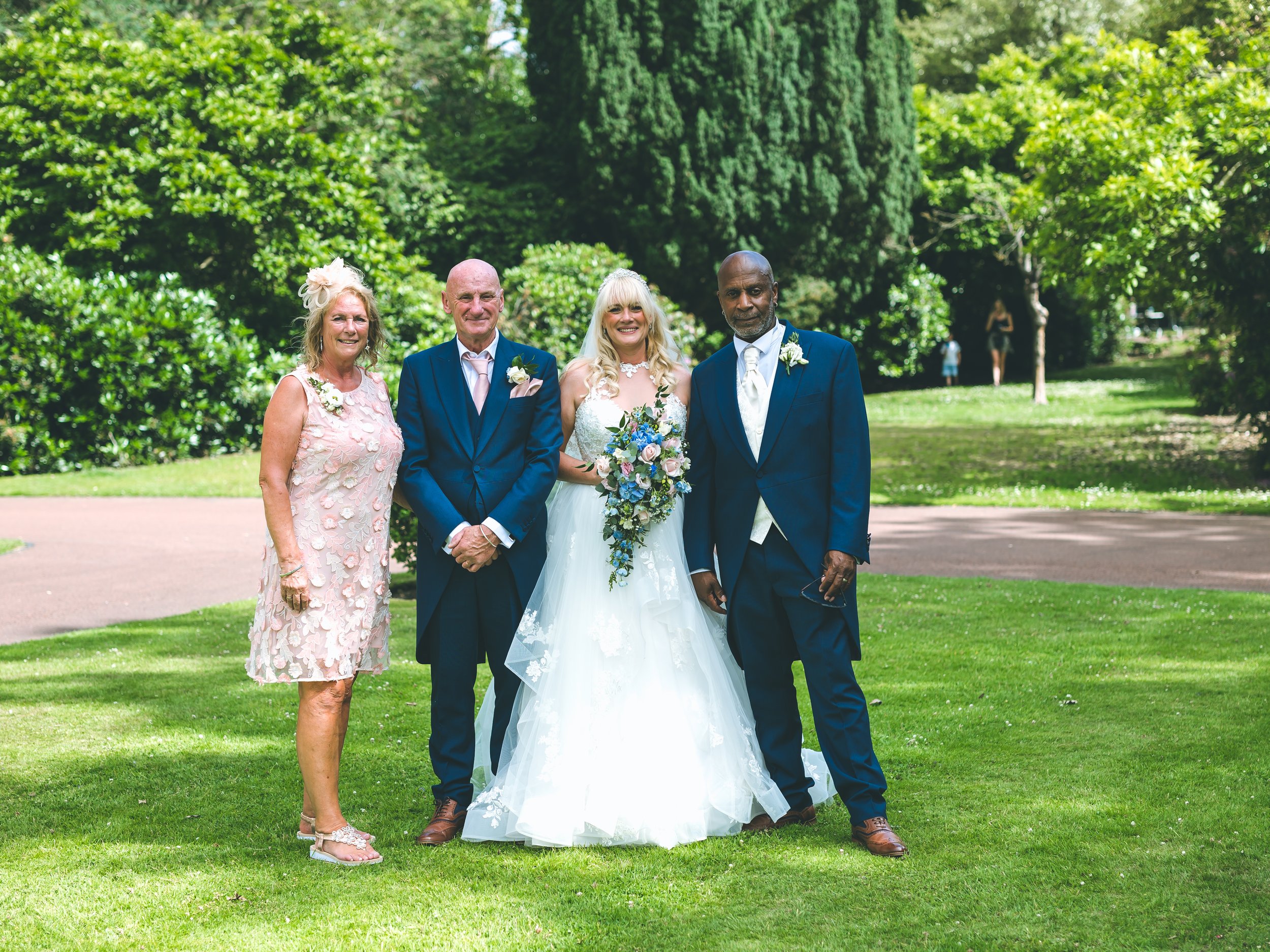 Four people standing outdoors on green grass in a park with trees in the background. The group includes a woman in a pink dress, two men in suits, and a woman in a white wedding dress holding a bouquet. The scene appears to be from a wedding or speci