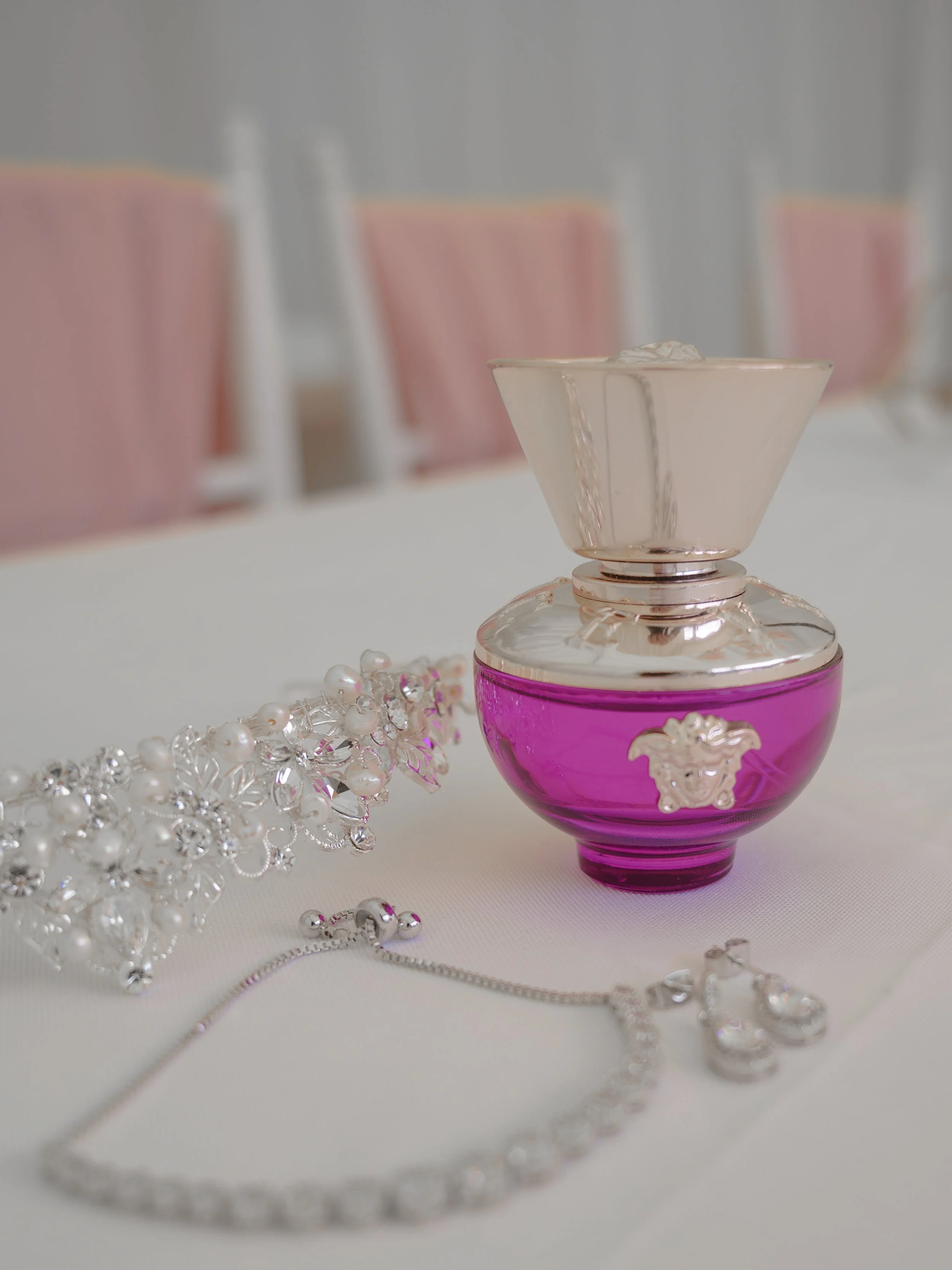 Close-up of a pink perfume bottle with a decorative clasp, surrounded by pearl and rhinestone jewelry on a white surface.