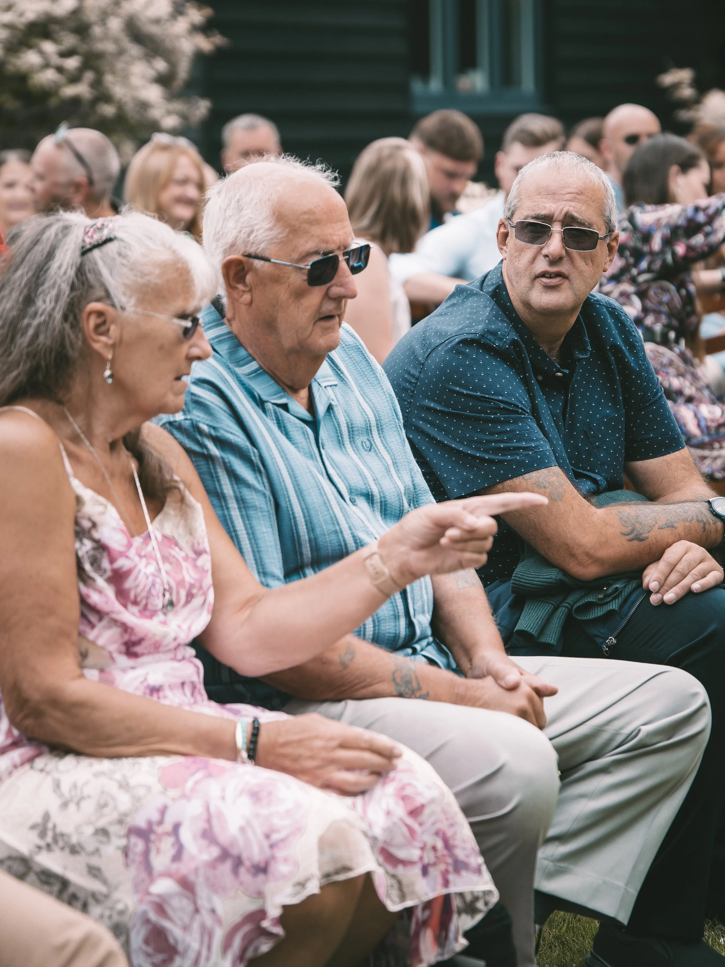 A group of elderly and middle-aged people sitting outdoors during daytime, some wearing sunglasses, and engaging in conversation or listening, in front of a dark green building with trees in the background.