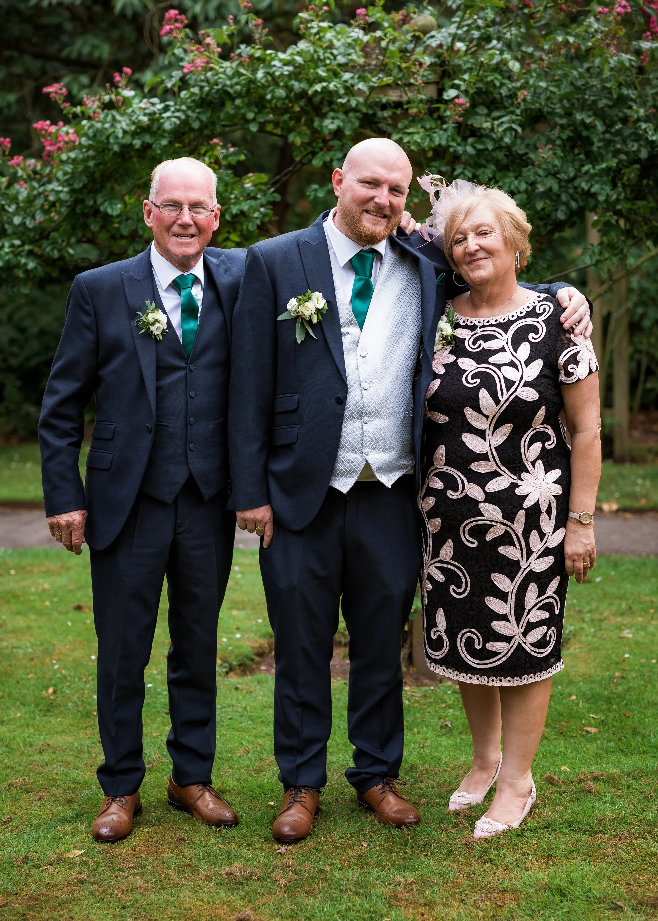 Four people standing on grass in front of pink flowering bushes, dressed in formal attire for a celebration or wedding, smiling and posing for a photo.