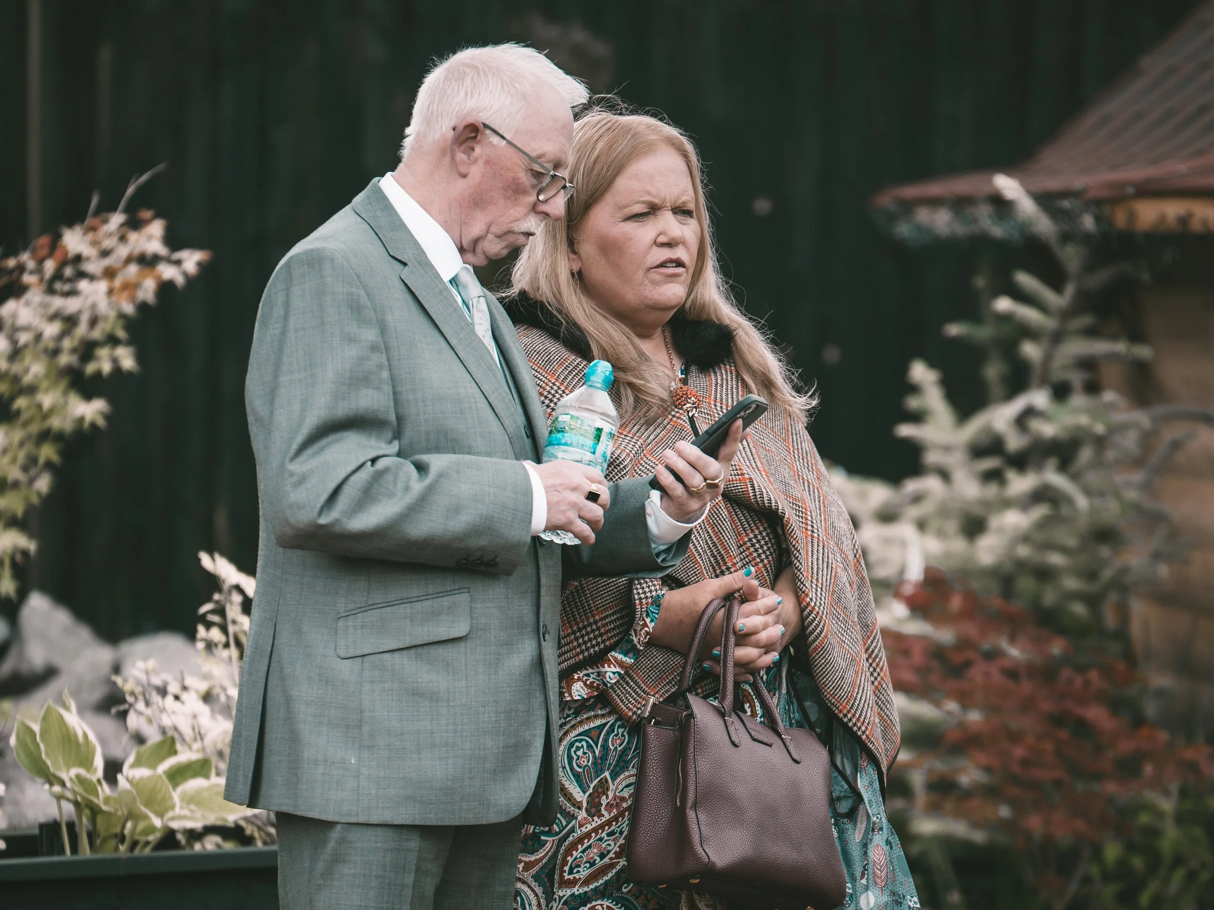 An elderly man and woman standing outdoors, looking at a smartphone together. The man is wearing a light gray suit and glasses, holding a water bottle. The woman has long, light brown hair and is wearing a patterned coat and holding a brown purse. Th