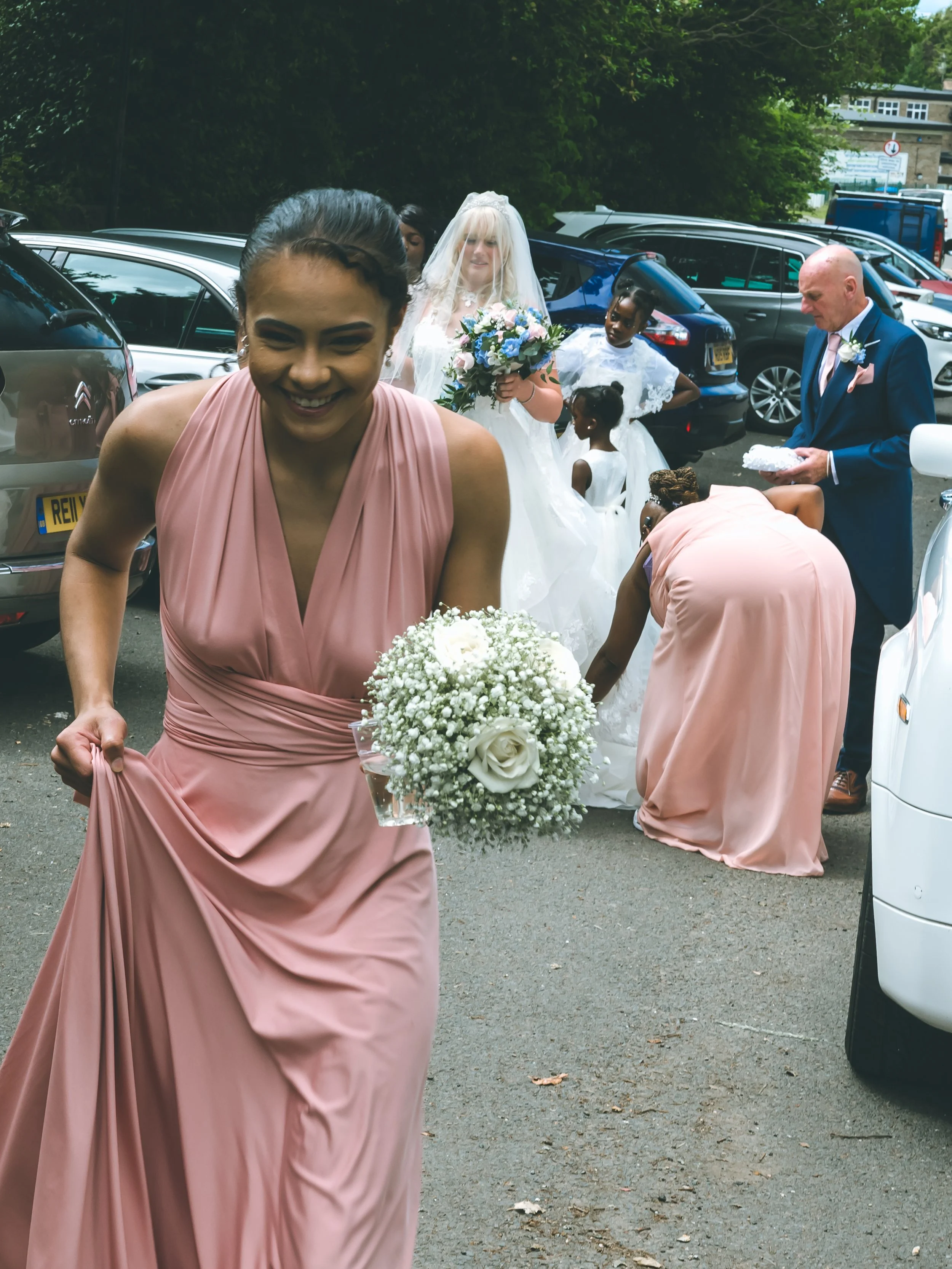 A bride in a white dress with a veil holding a bouquet of flowers, surrounded by bridesmaids in pink dresses, with men in suits, in a parking lot.