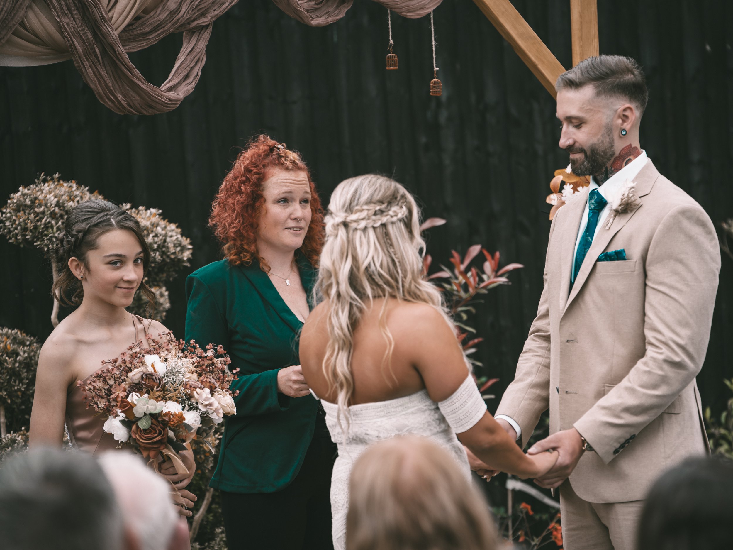 A wedding ceremony taking place outdoors with a woman in a white dress exchanging vows with a man in a beige suit, while a young girl with dark hair and a flower crown holds a bouquet nearby, and a woman with curly red hair watches the exchange.