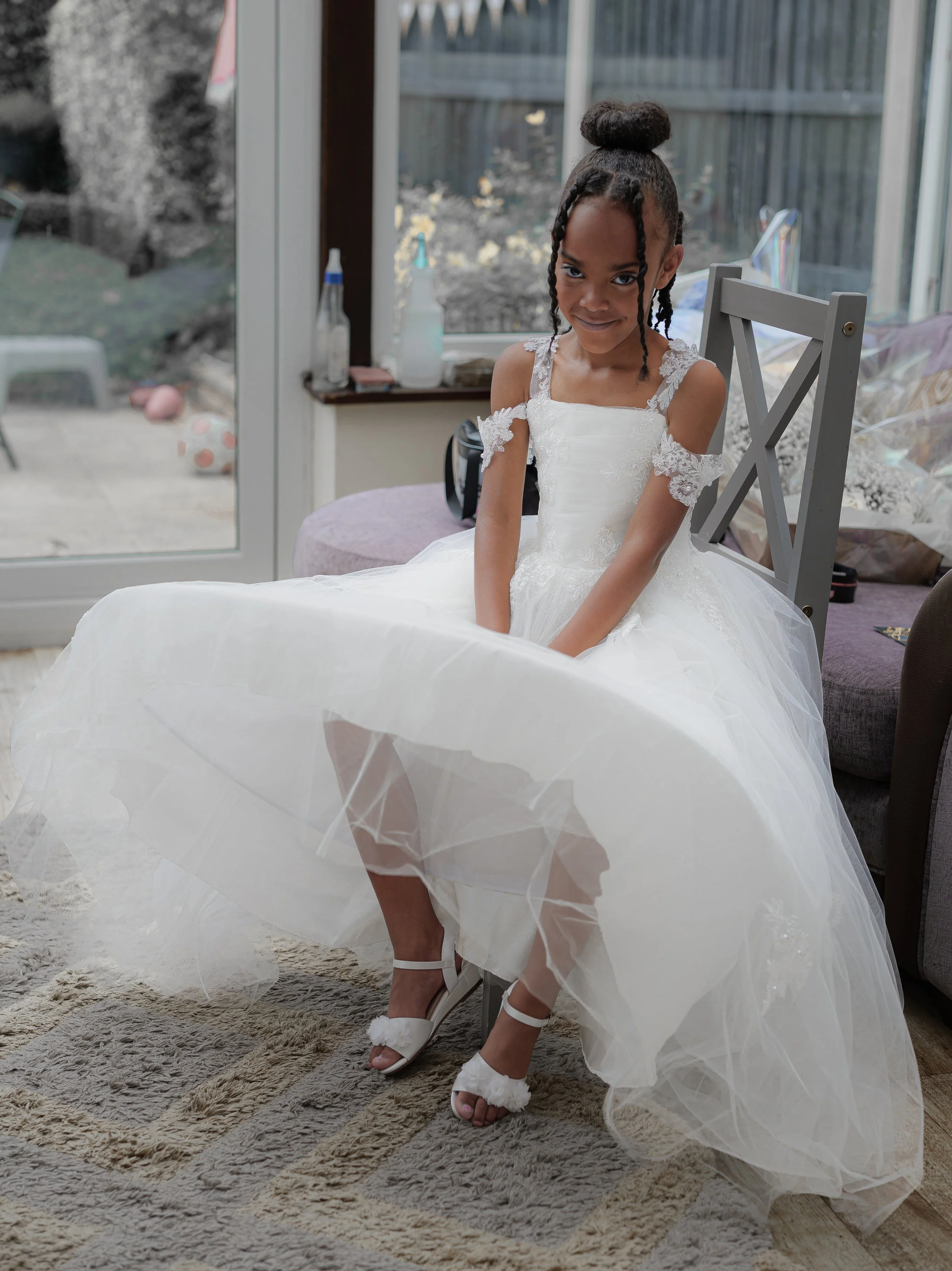 A young girl in a white dress sitting on a chair inside, with a sliding glass door behind her leading to a backyard. She is lifting her dress slightly, showing her white shoes with fluffy details.