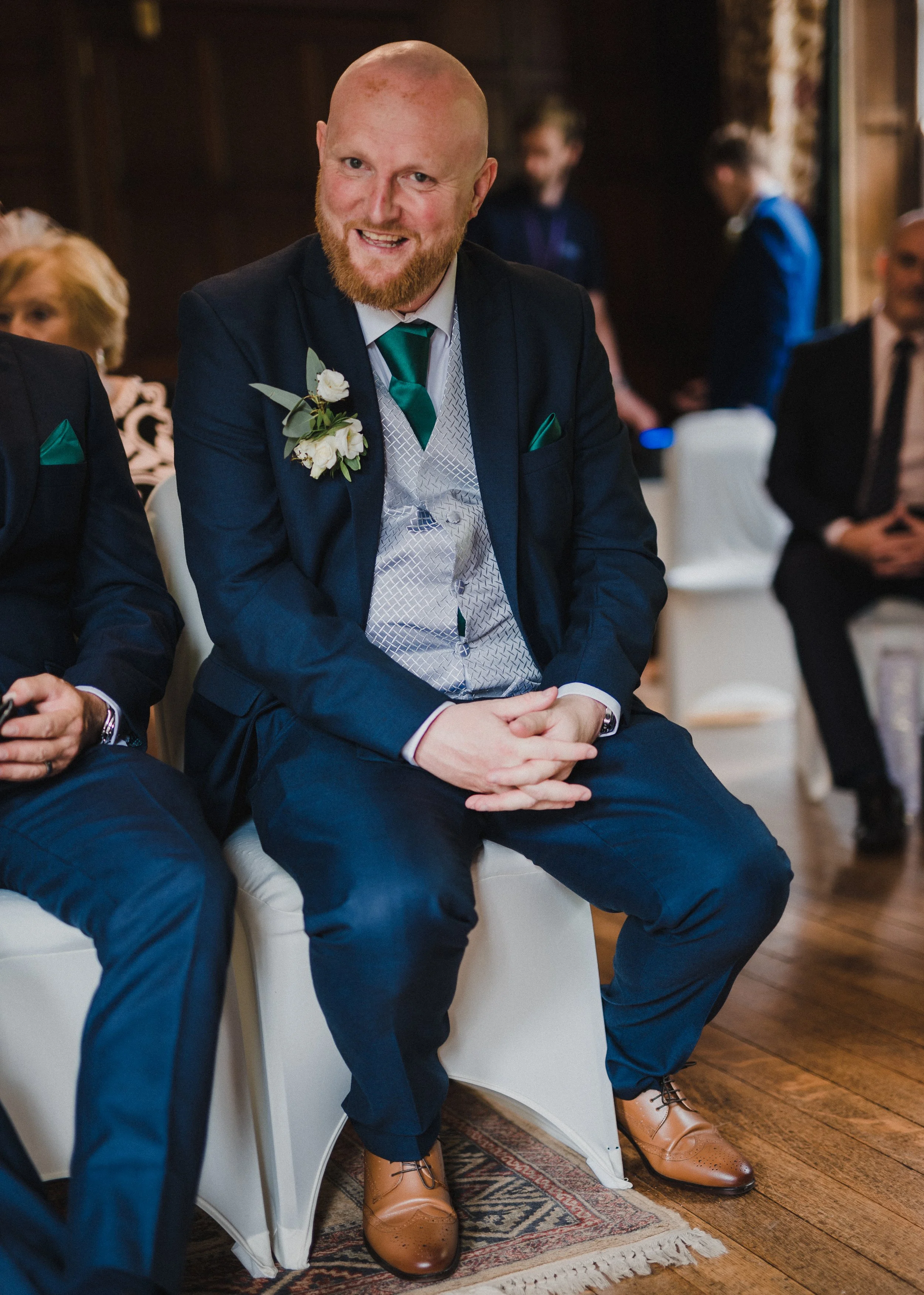 A bearded man in a blue suit with a white shirt and a green tie, wearing a boutonniere, sitting on a white chair at a formal event, smiling, with polished brown dress shoes, an ornate rug, and other people in formal attire in the background.