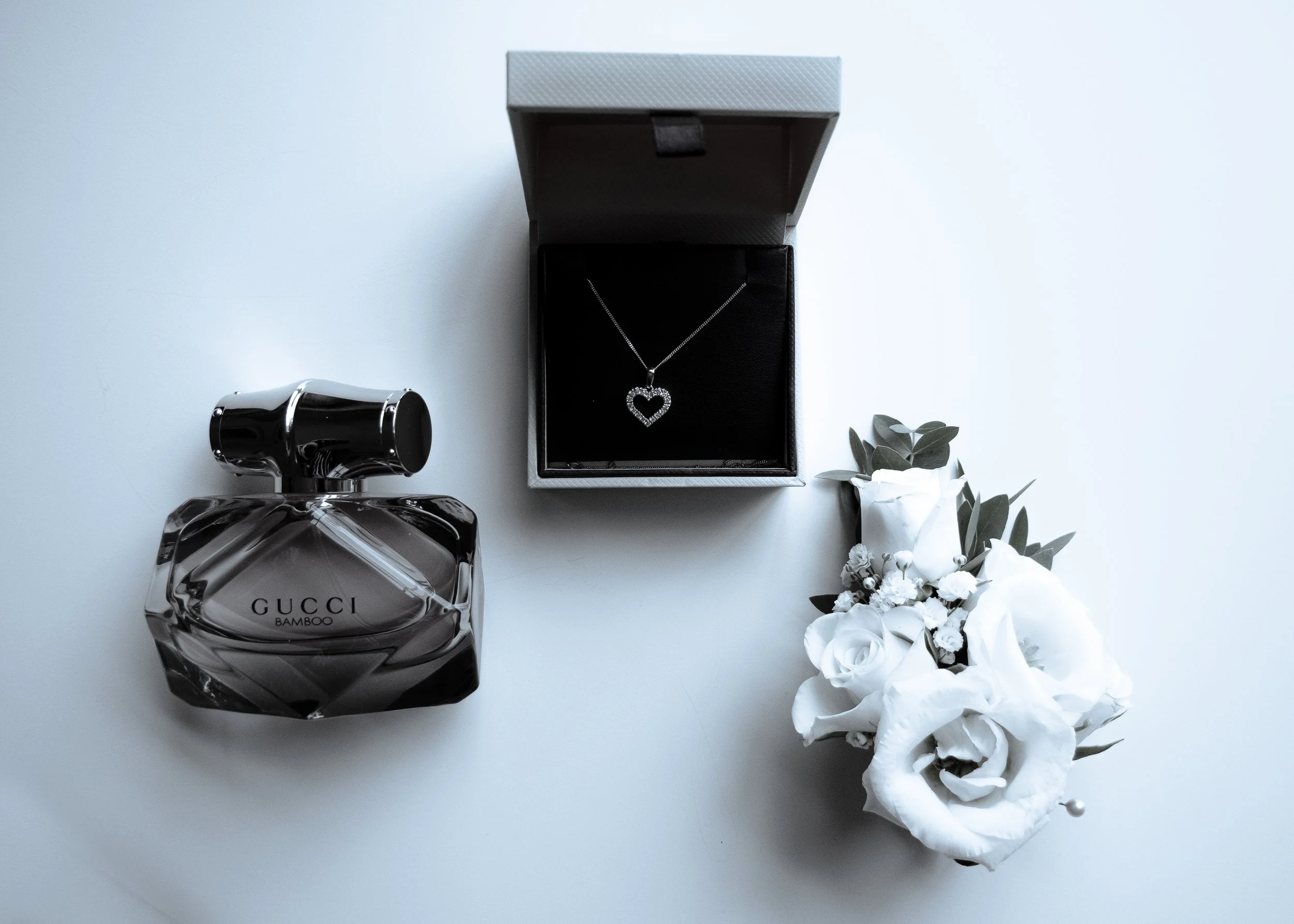 A black jewelry box with a necklace inside, a clear perfume bottle, and a bouquet of white flowers with green leaves.