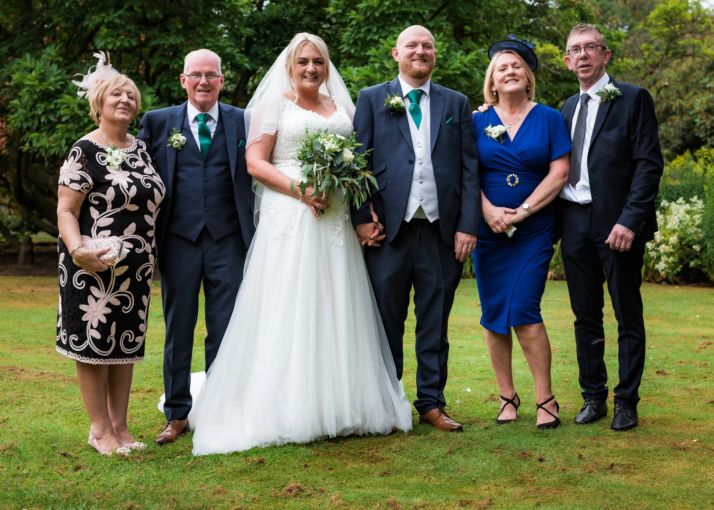 Group of six people, including a bride in a white wedding dress holding a bouquet, and five other individuals, standing outdoors on grass with trees in background, smiling for the camera.