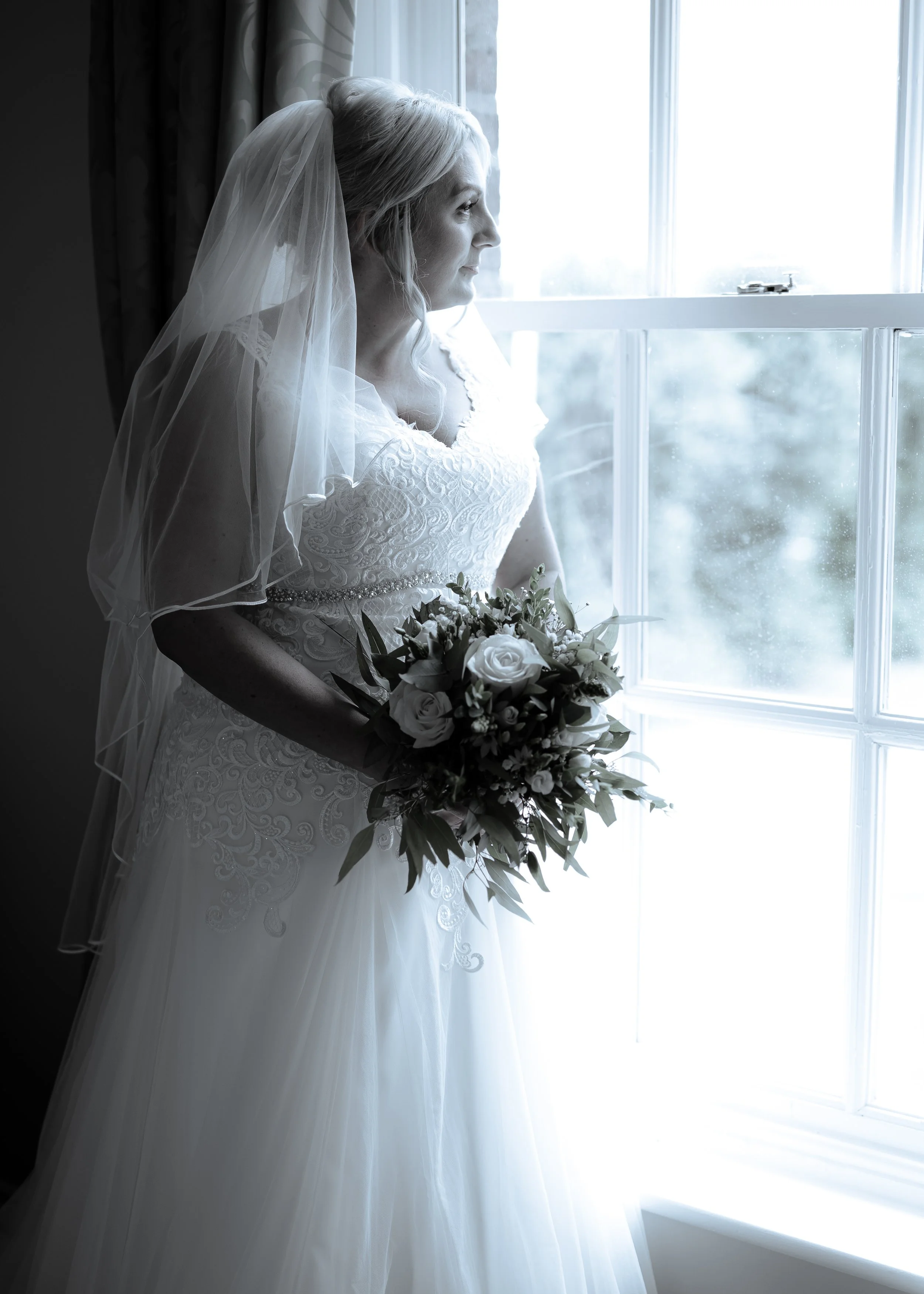 A bride in a white lace wedding gown with a veil holding a bouquet of white and green flowers, standing by a window and looking outside.