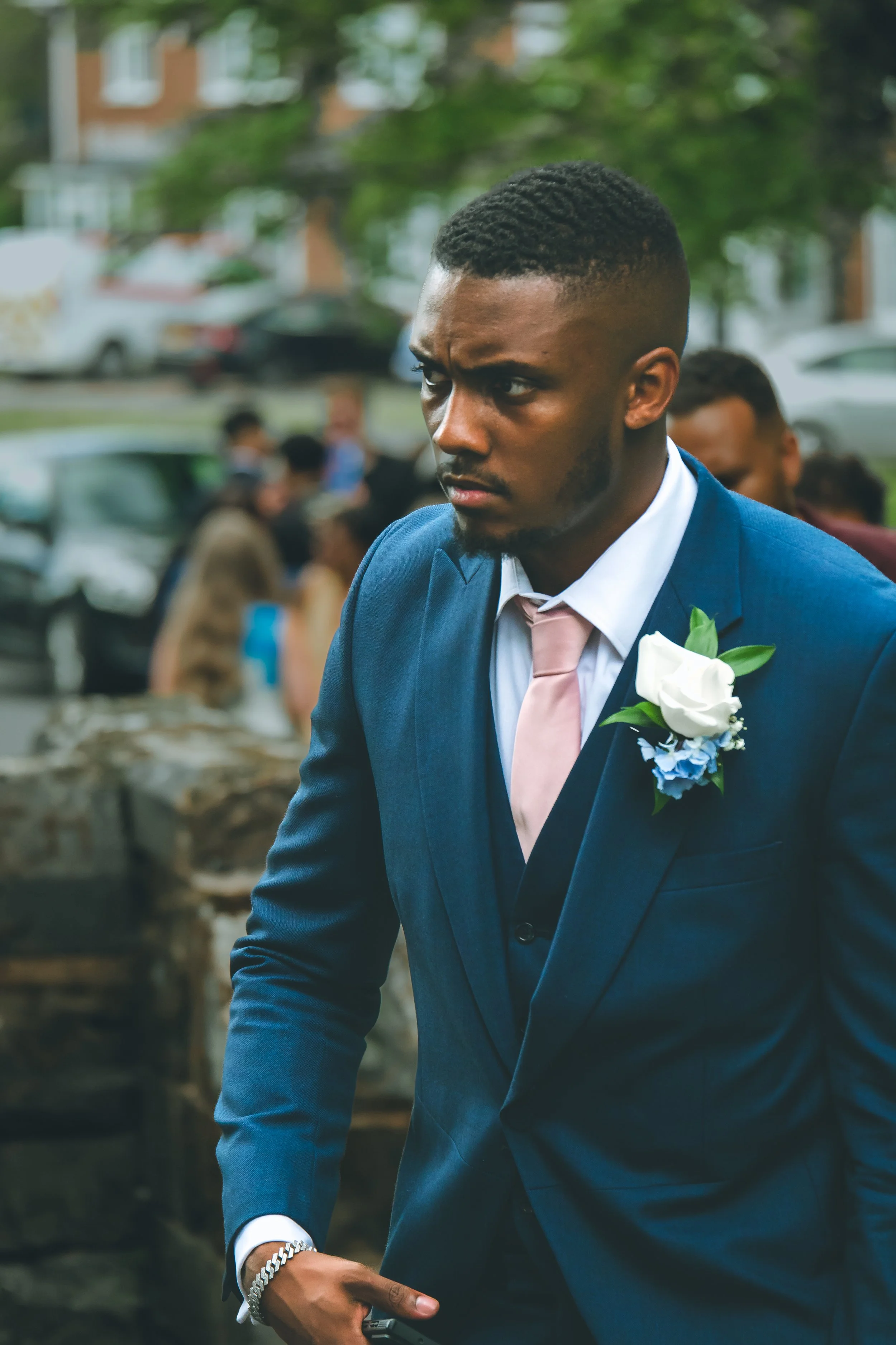 A young man dressed in a blue suit with a white shirt and pale pink tie, wearing a white boutonniere with blue flowers, standing outdoors with a serious expression on his face. There are blurred people and cars in the background.