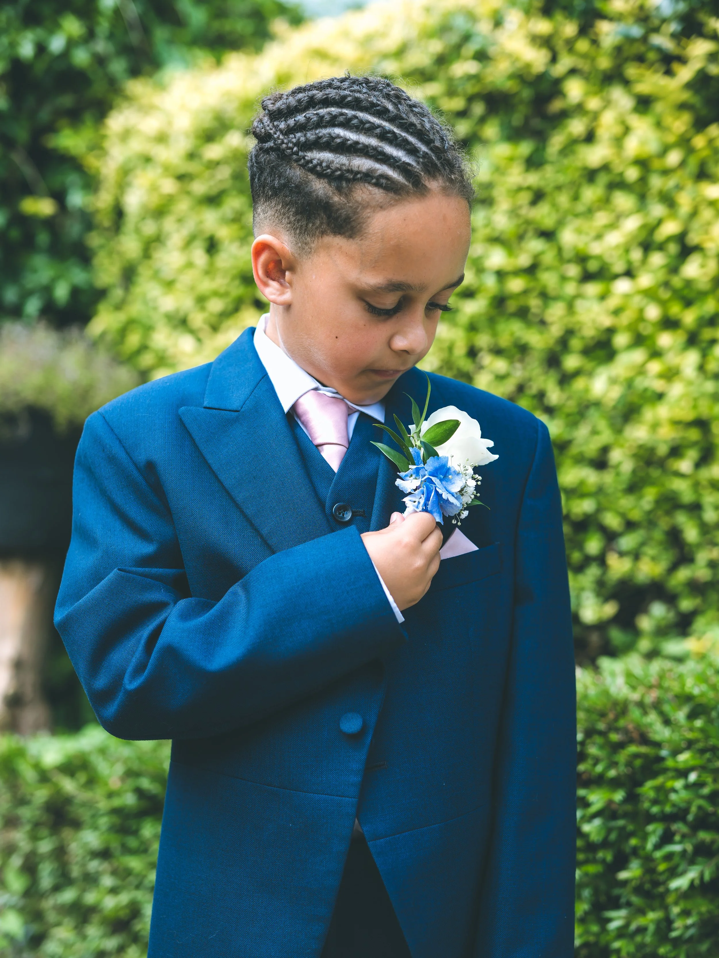 A young boy dressed in a navy blue suit with a pink tie, holding a small bouquet of white and blue flowers, standing outdoors with a blurred green natural background. He is looking down and gently touching his chest.