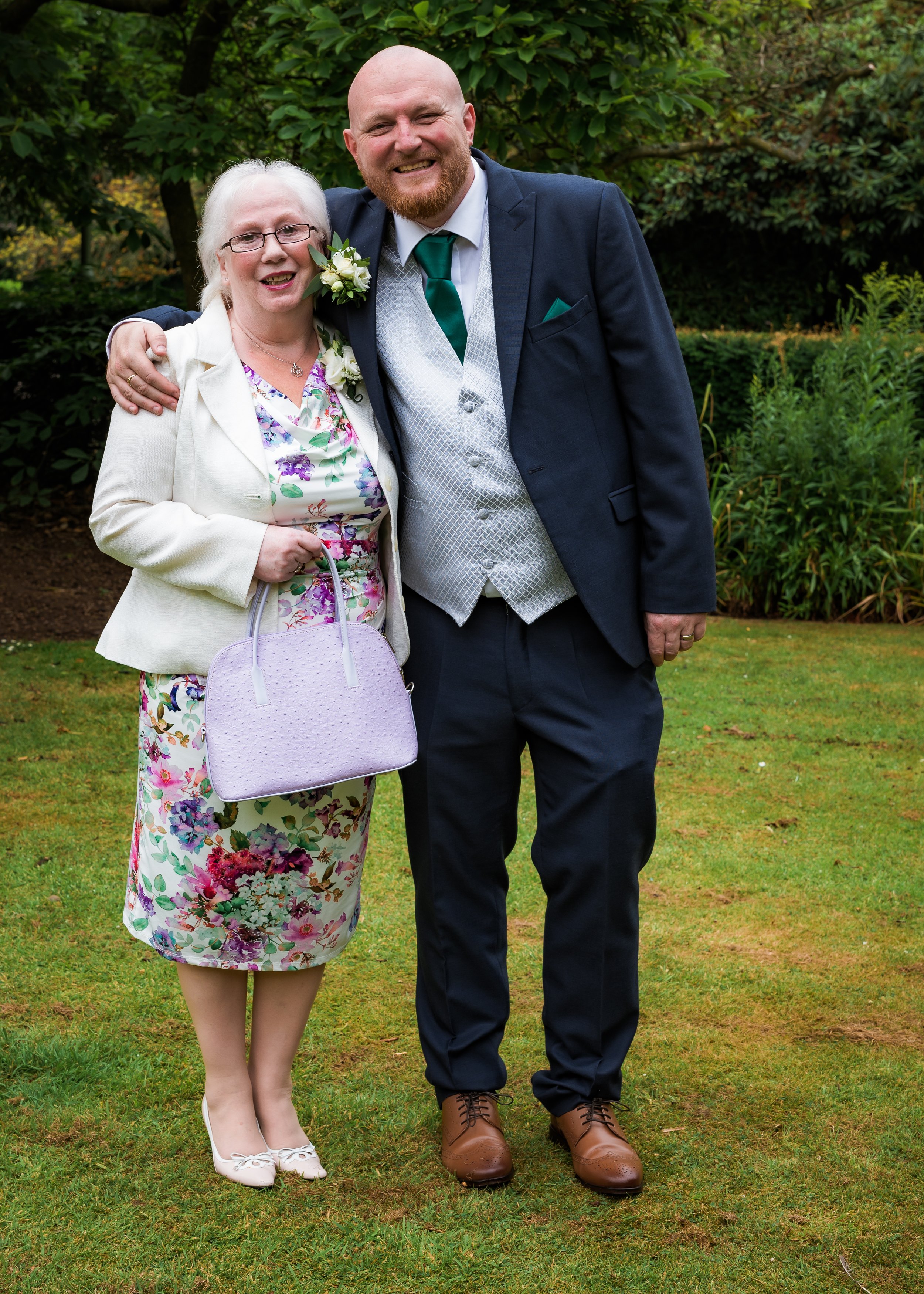 Senior woman and man dressed in formal attire standing outdoors on grass with trees in the background, smiling and posing for the photo.