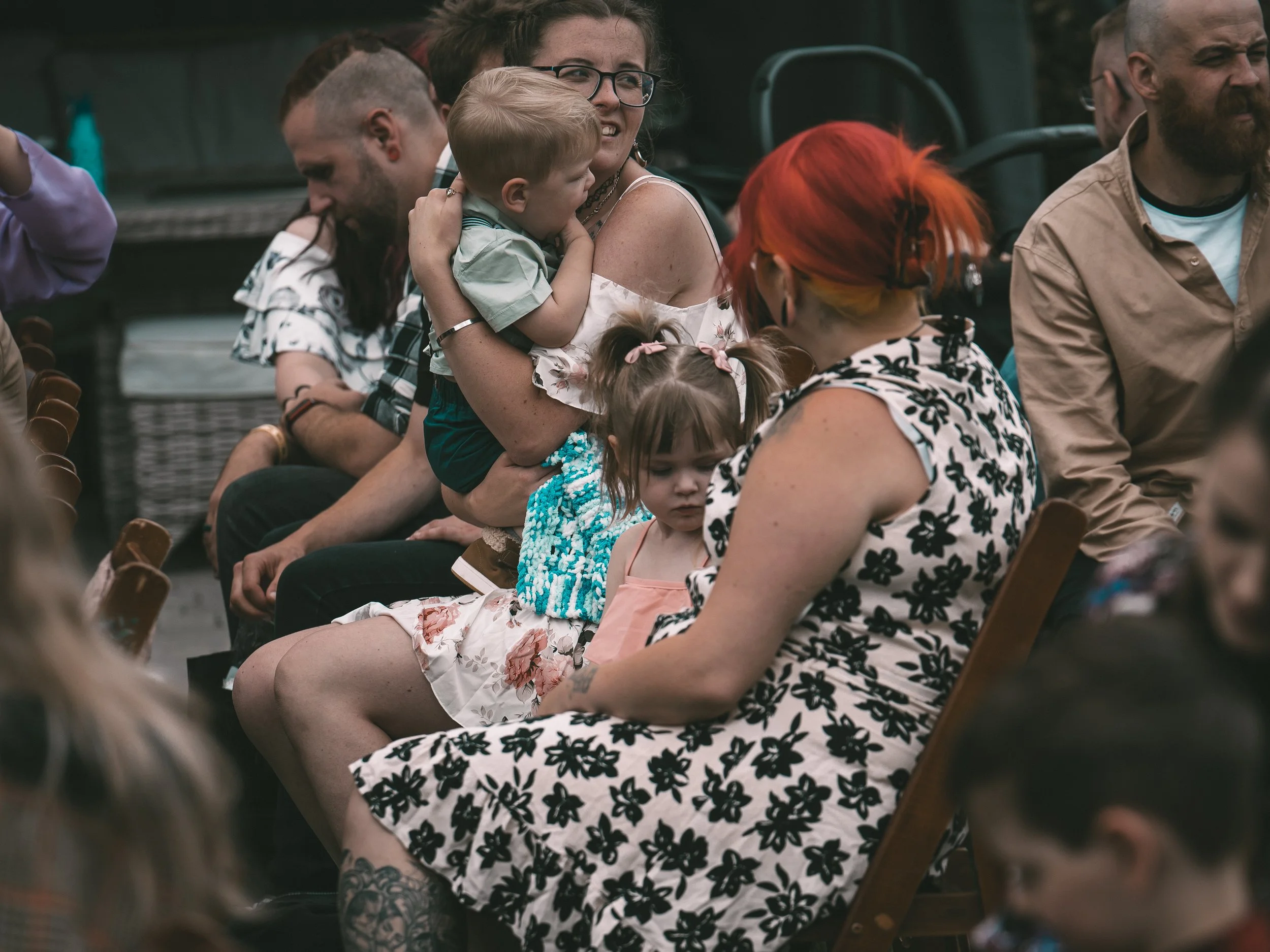 A group of people sitting in a row, with one woman holding a young child in her lap, who appears to be asleep, at an outdoor event during the daytime.