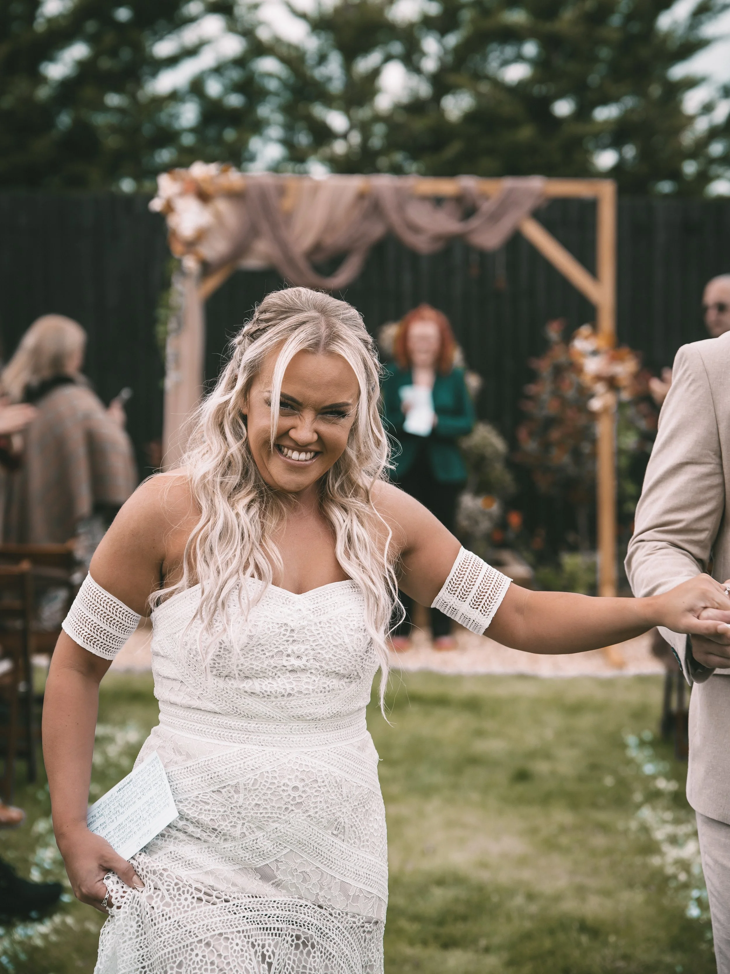 A smiling blonde woman in a white lace dress at an outdoor wedding ceremony, with a black fence and trees in the background, and a wooden arch decorated with flowers and fabric.