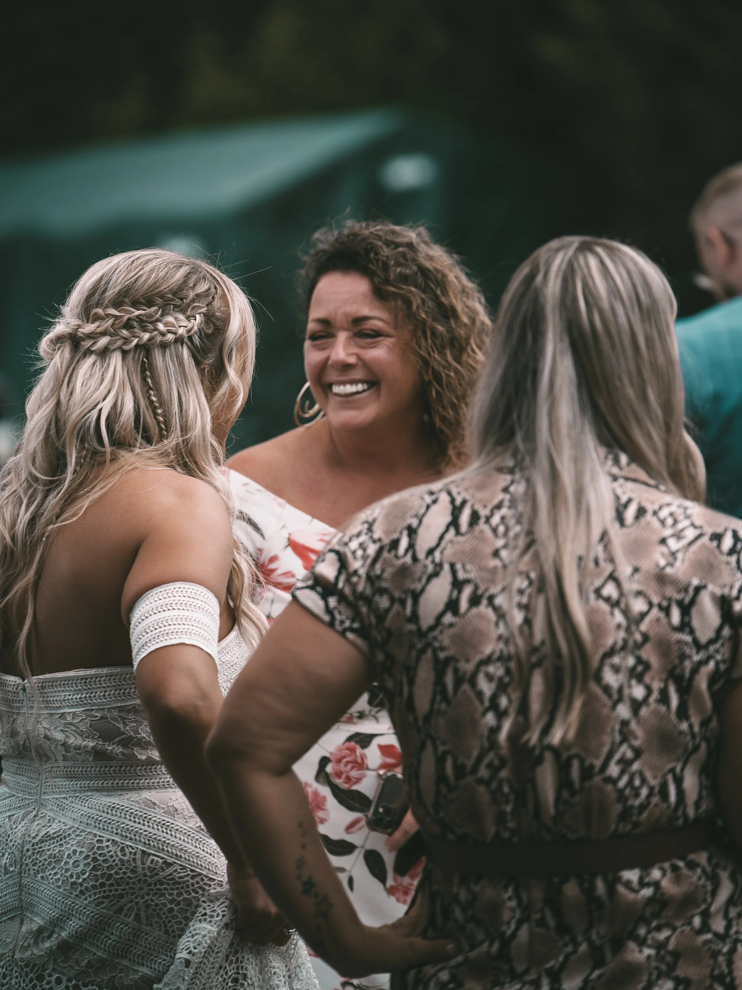 A group of four women engaged in conversation outdoors, smiling and laughing. One woman has long blonde hair styled in braids and wears a white lace dress. The others have curly and straight hair and wear patterned clothing.