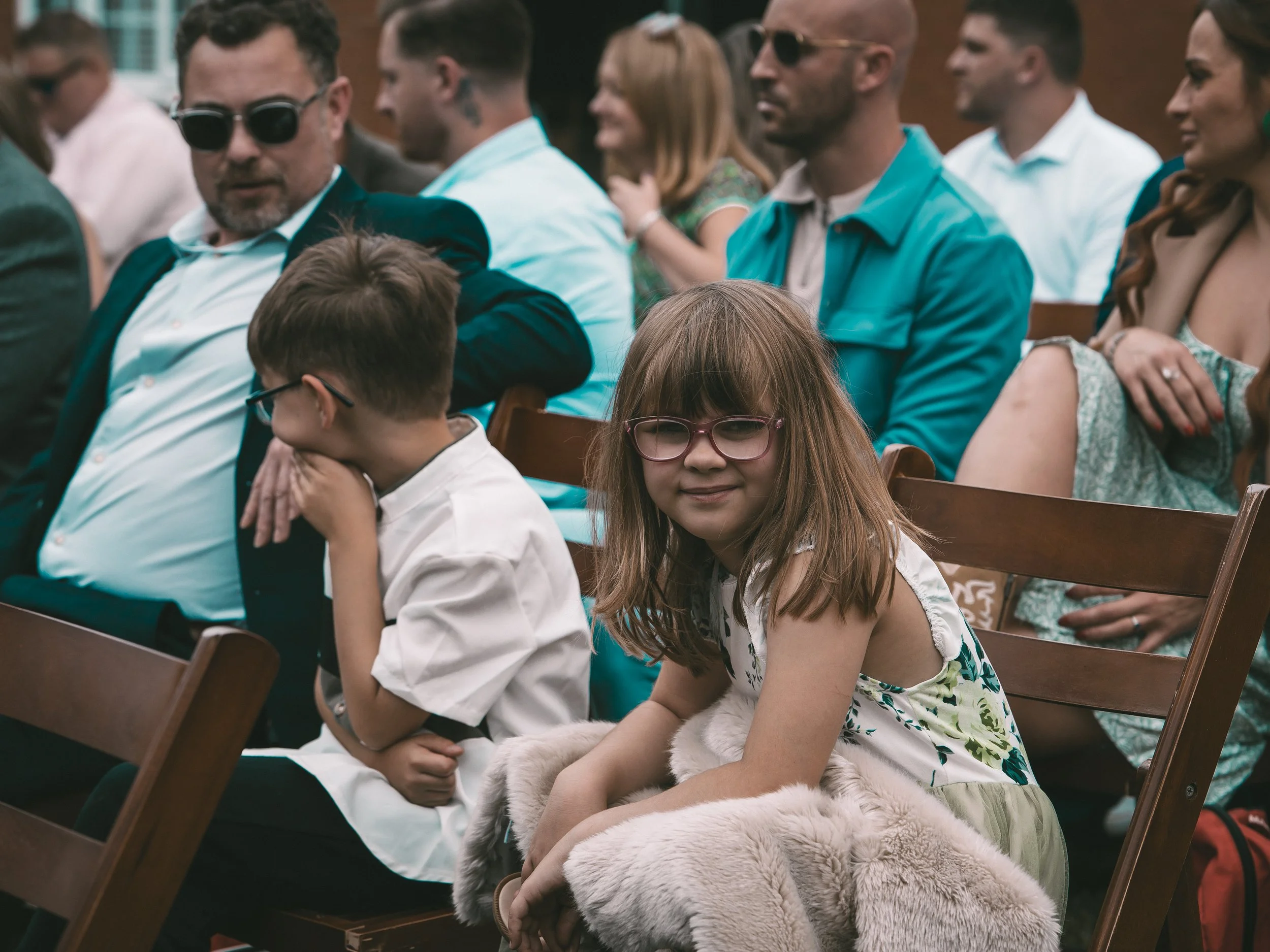 People seated in chairs attending an outdoor event, with some socially distanced and dressed in formal clothing.