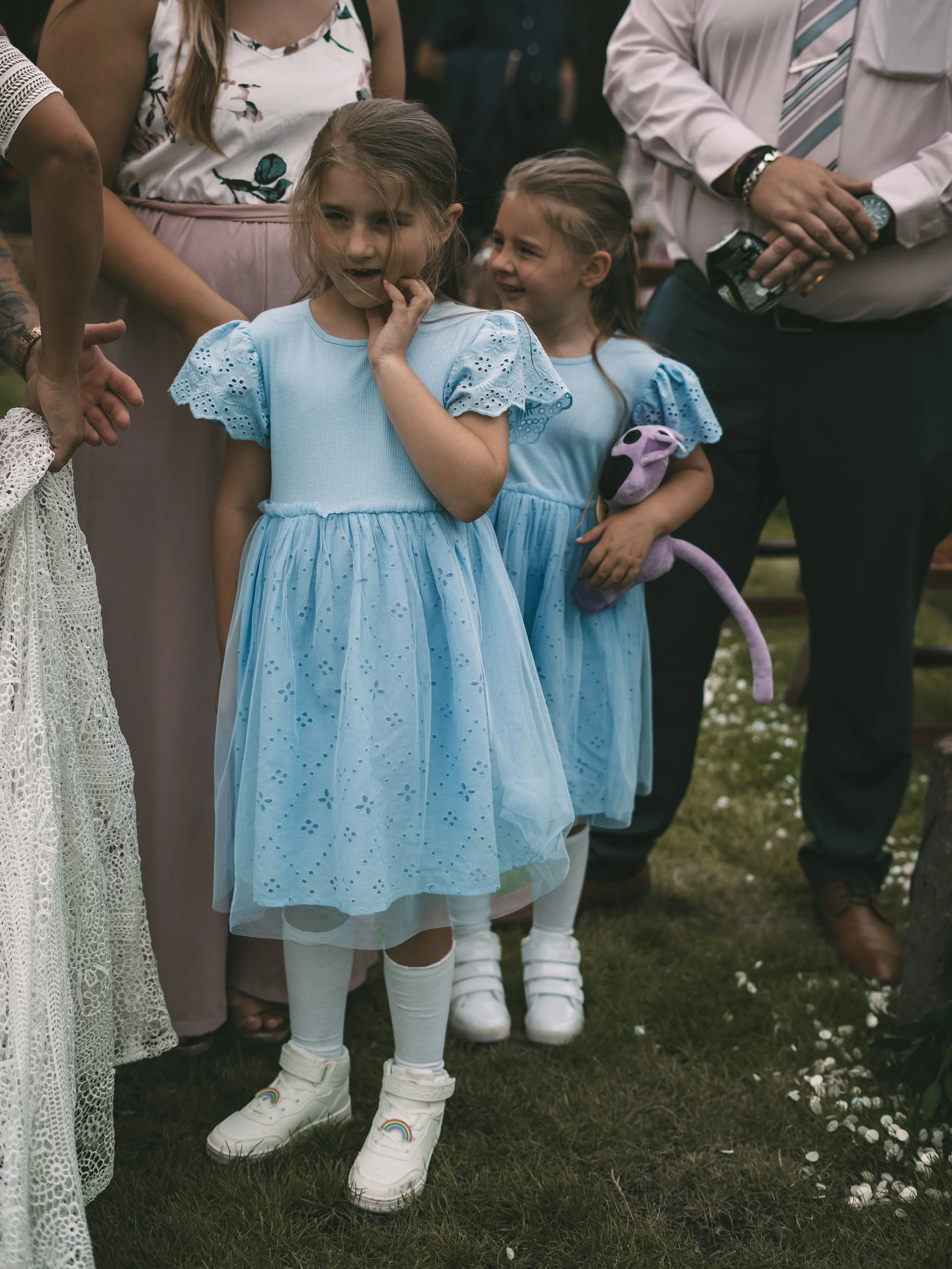 Two young girls dressed in matching light blue dresses with puffed sleeves and eyelet details, wearing white tights and sneakers with rainbow designs, standing outdoors. Two adults, a woman and a man, are partially visible around them, with the woman