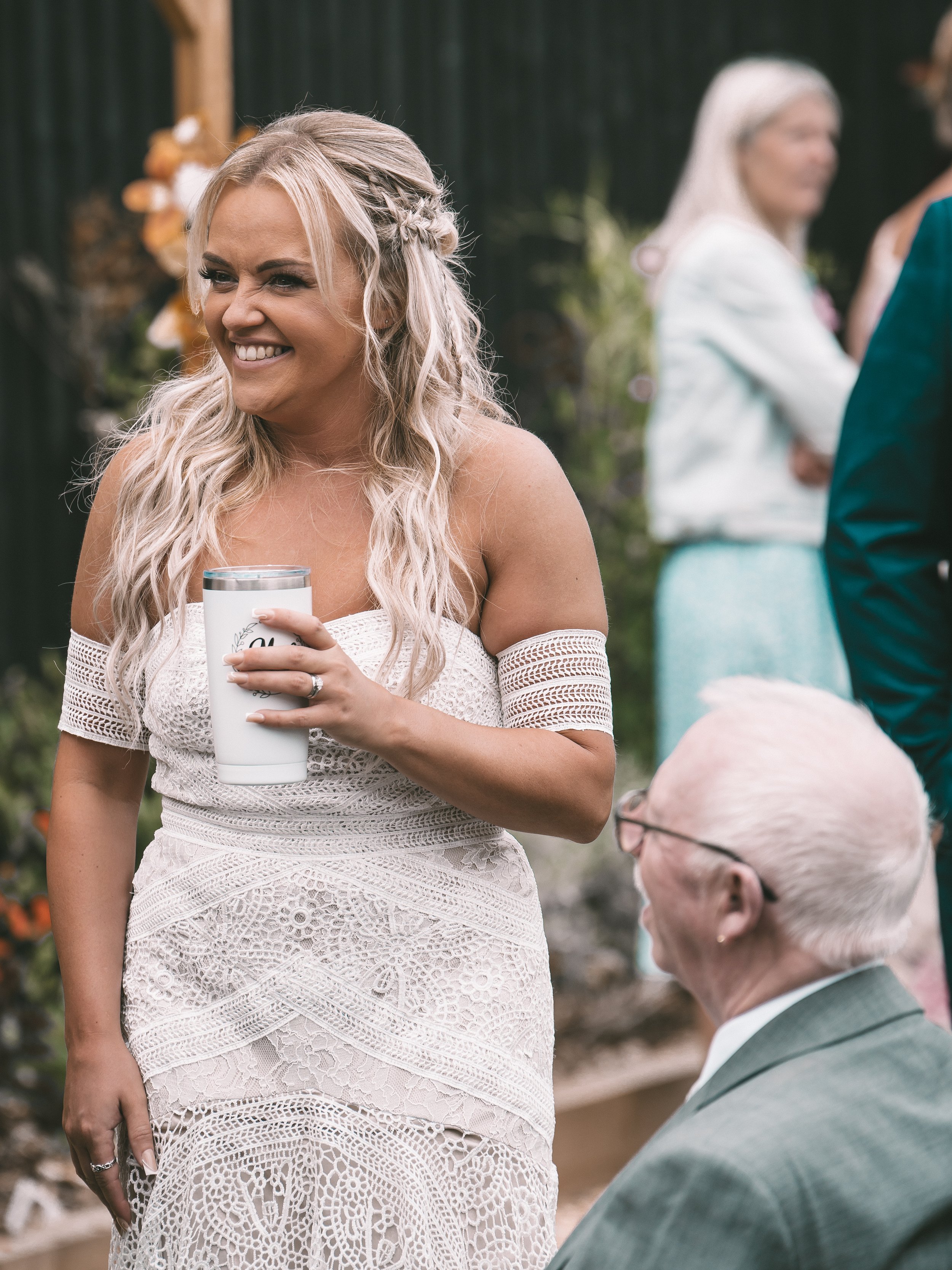 A smiling bride with long blonde hair styled in loose curls and a small braid, wearing a white lace dress with off-the-shoulder sleeves, holding a white tumbler cup, at an outdoor wedding with three guests in the background.