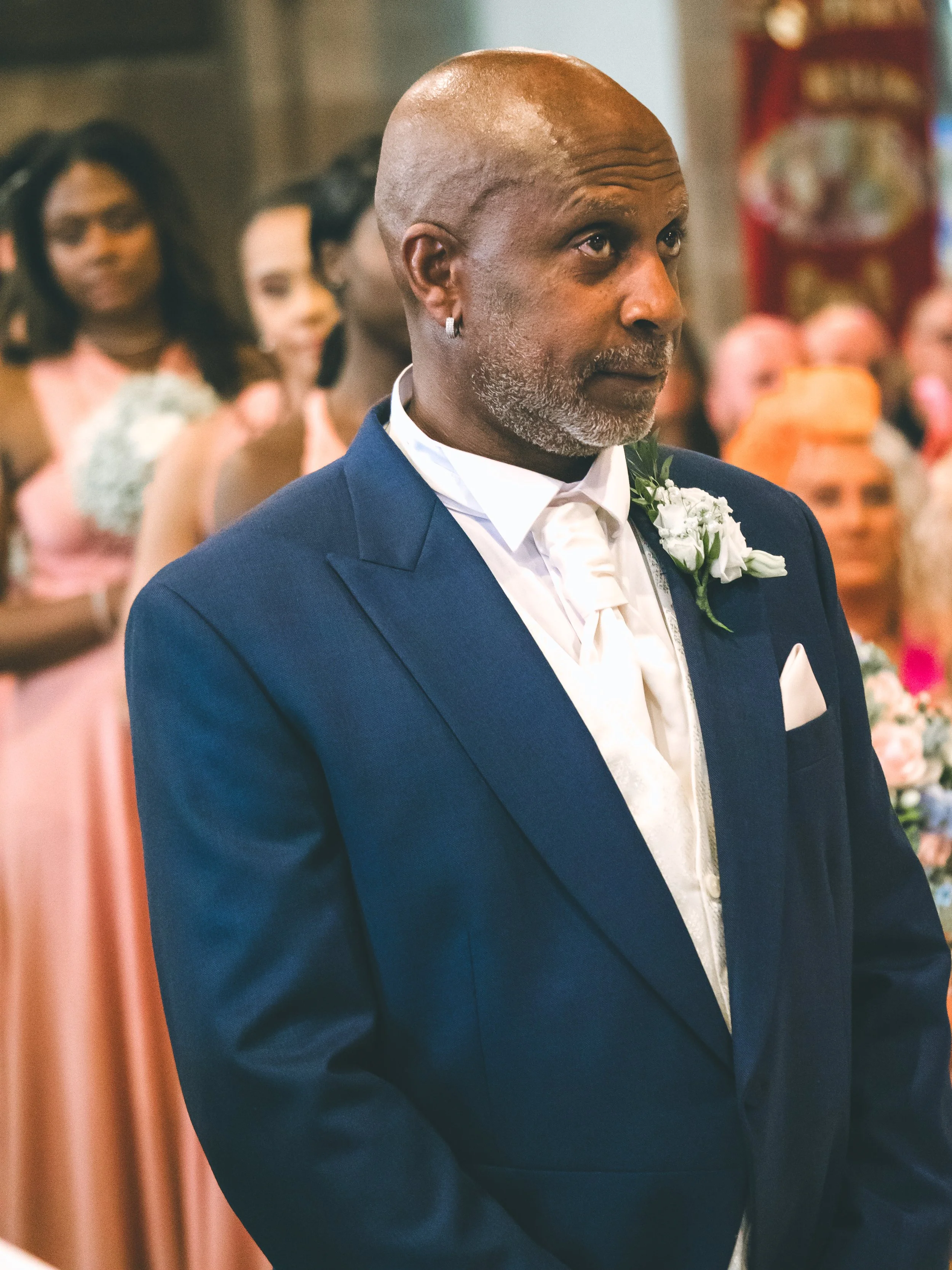 Close-up of a man with a mixed gray and bald head, wearing a blue suit and white shirt with a boutonnière, standing indoors with blurred people in the background.