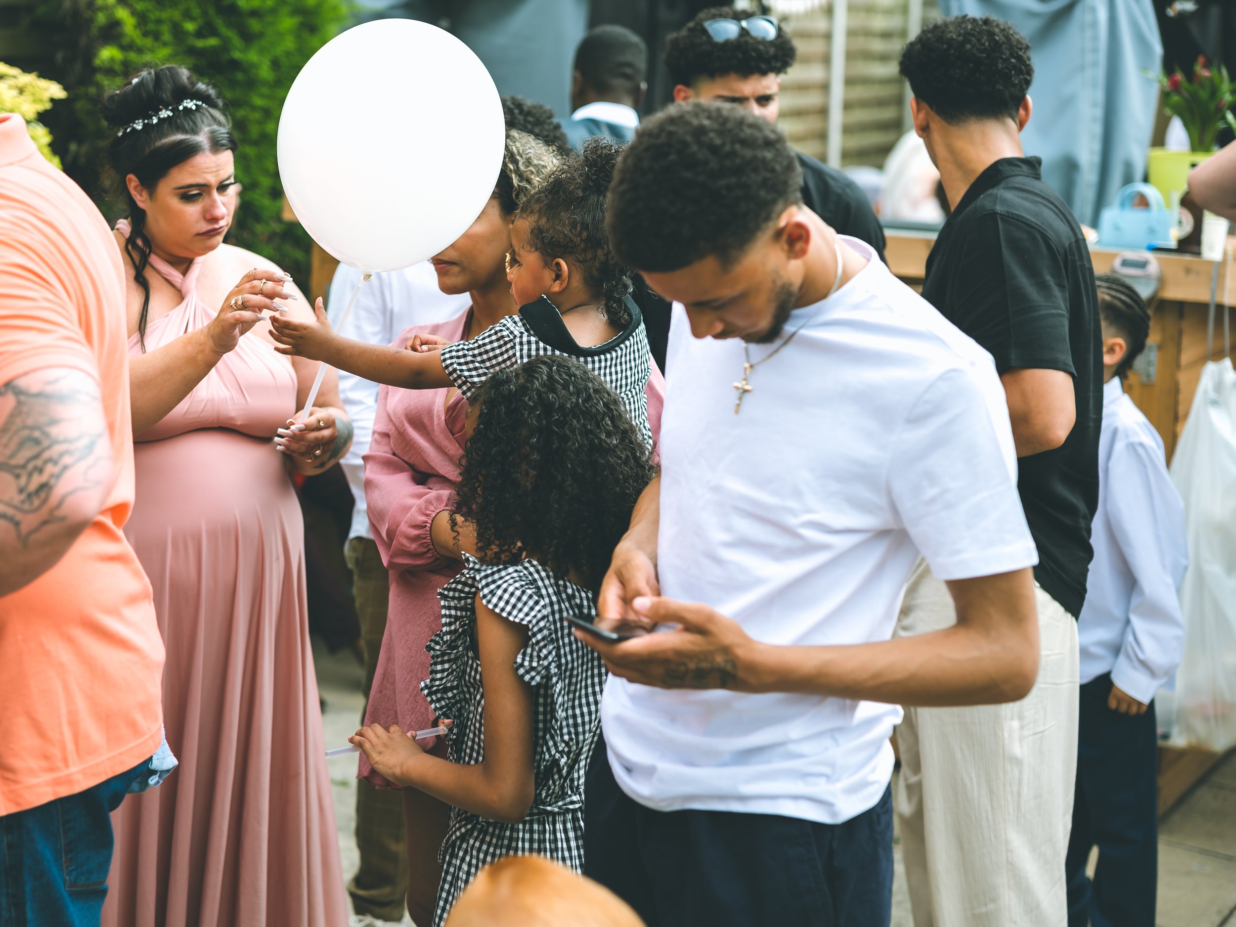 A group of people, including children and adults, gather outdoors at a daytime event. Some are looking at their phones, others are engaging with each other. There are colorful decorations and tables in the background, suggesting a social or family ga