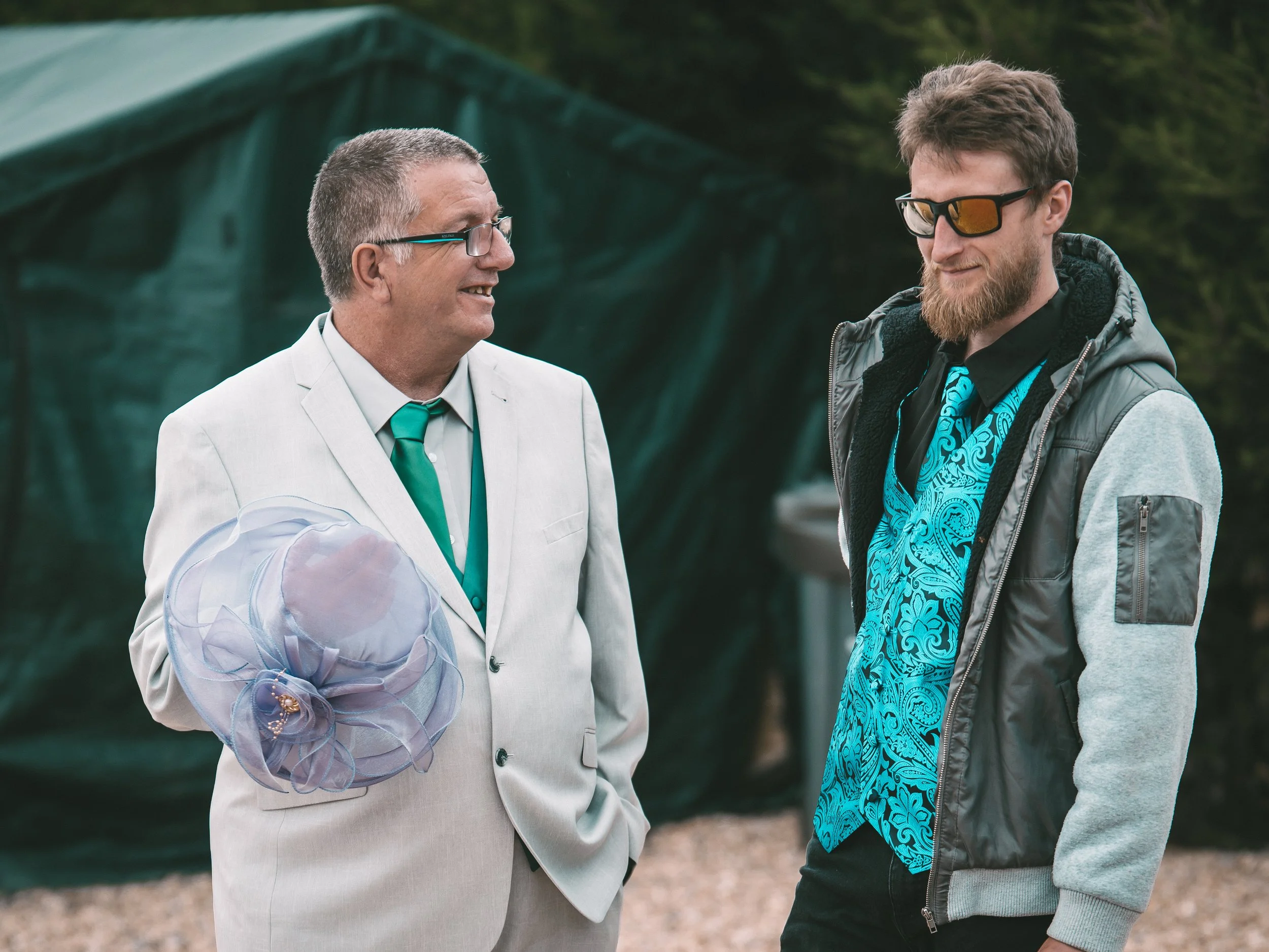 Two men talking outdoors, one in a light gray suit with a teal tie and the other in a gray hoodie with sunglasses, standing in front of a green tent and trees.