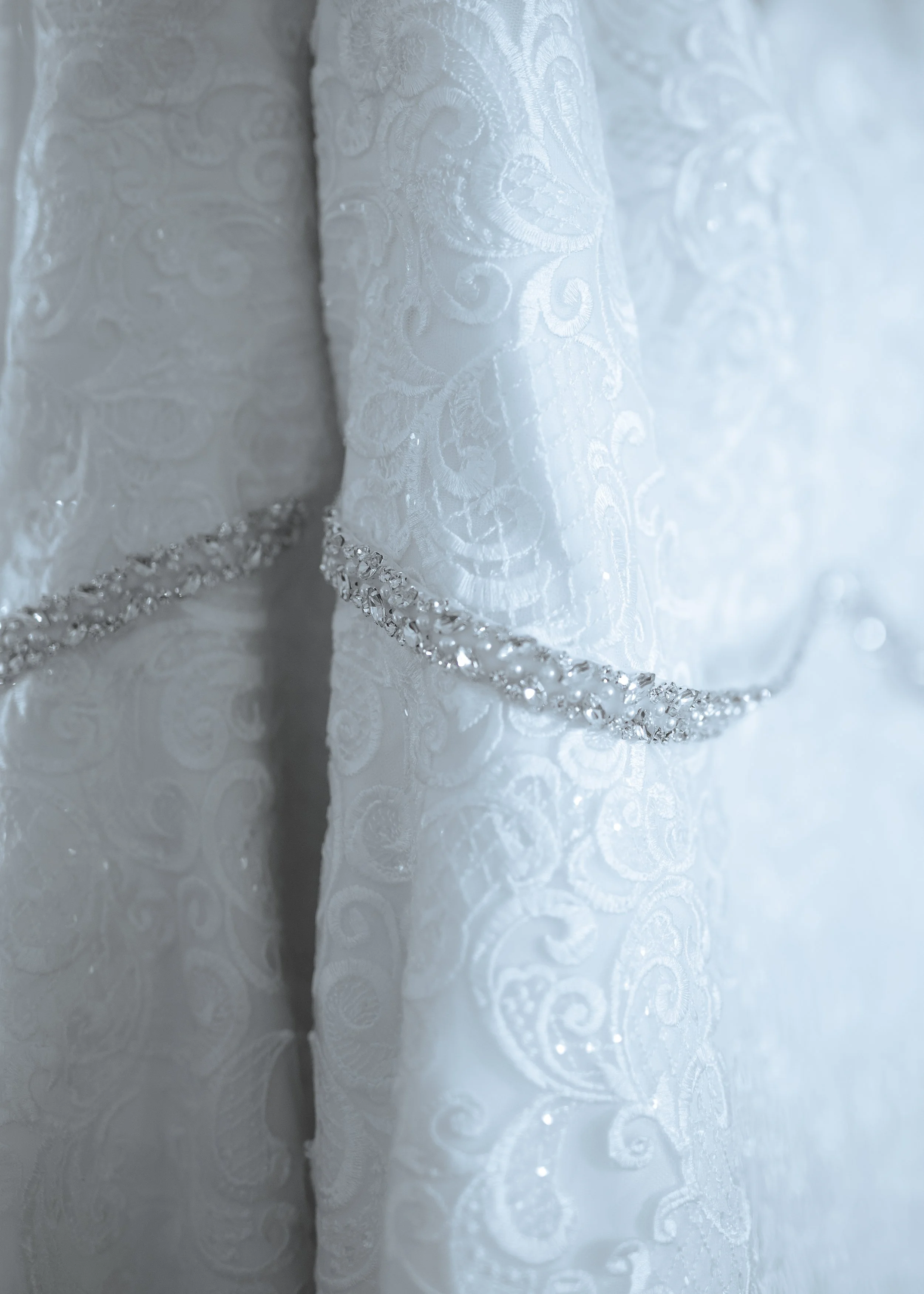 Close-up of luxurious white fabric with intricate swirling patterns and a decorative border of rhinestones or pearls.
