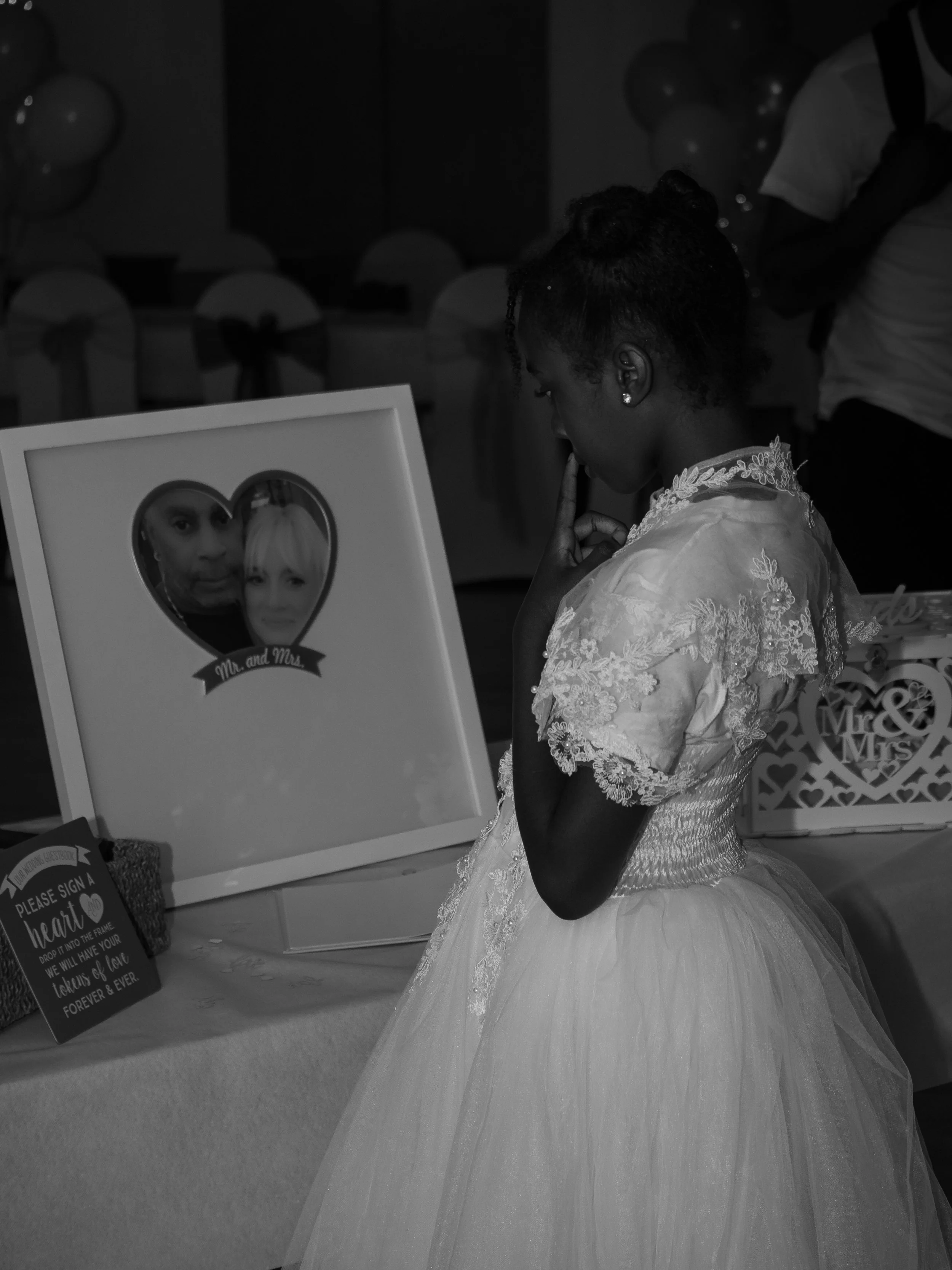A woman in a white lace wedding dress, with her hair styled in an updo, is standing at a table looking at a framed photograph of a couple, with her finger to her lips in a contemplative gesture. The scene appears to be at a wedding or reception, with