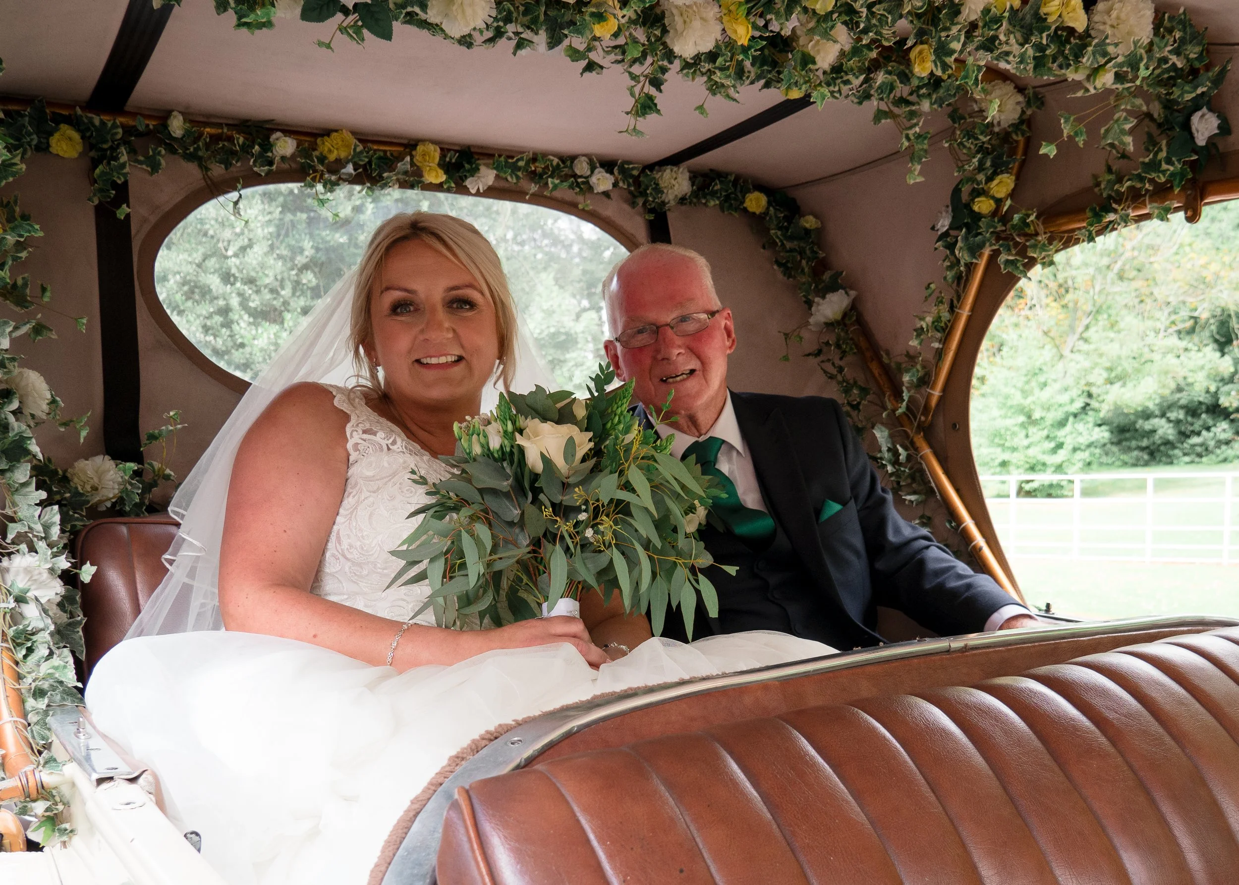 A bride and an older man, likely her father, sitting inside a decorated carriage. The carriage is adorned with green and white flowers and ivy. The bride is wearing a white wedding dress and veil, holding a bouquet of white roses and greenery, and ha