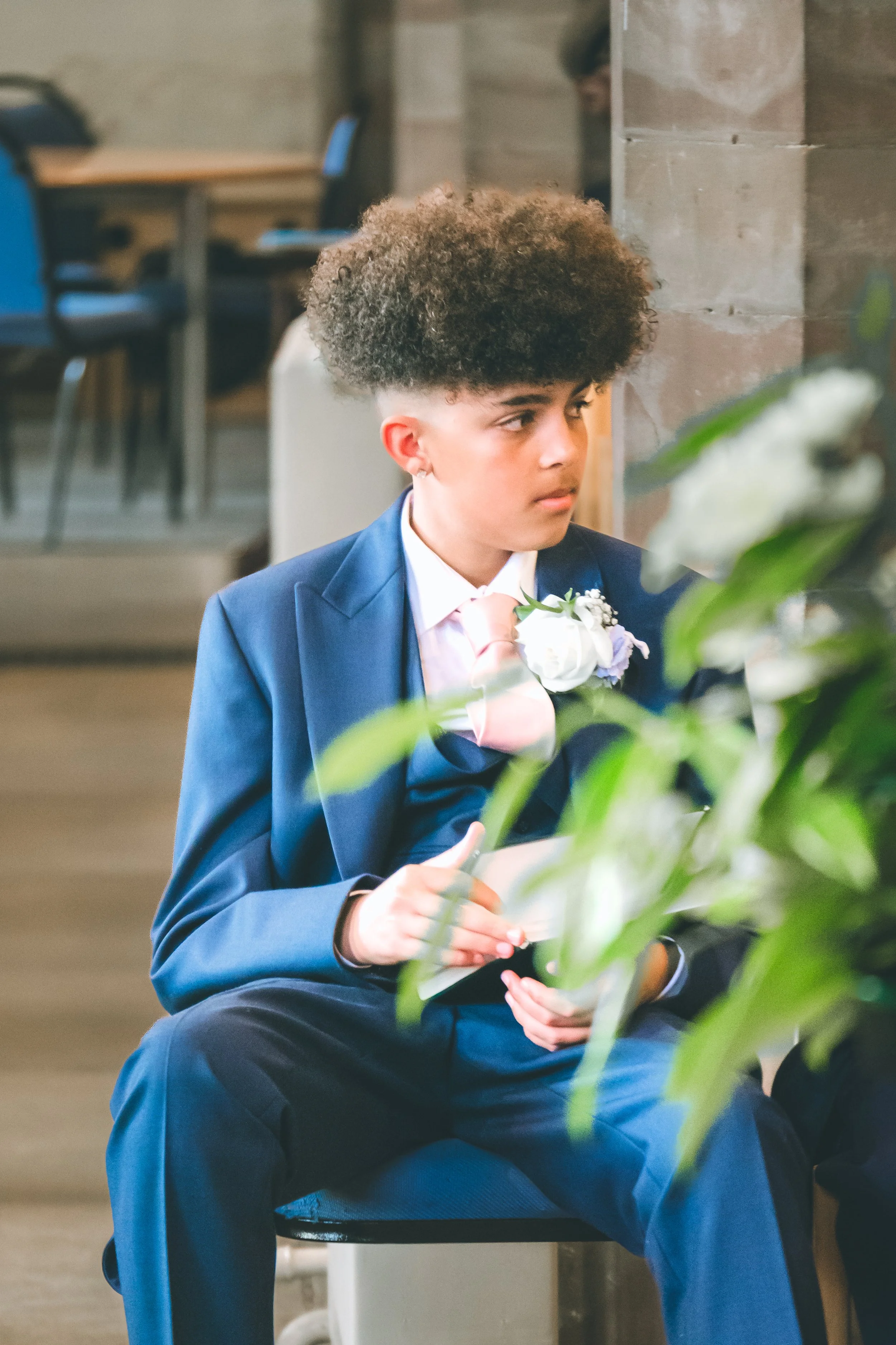A person with curly hair wearing a dark blue suit and a light pink tie, sitting on a chair indoors, holding a piece of paper, with a blurred background of chairs and a stone wall.