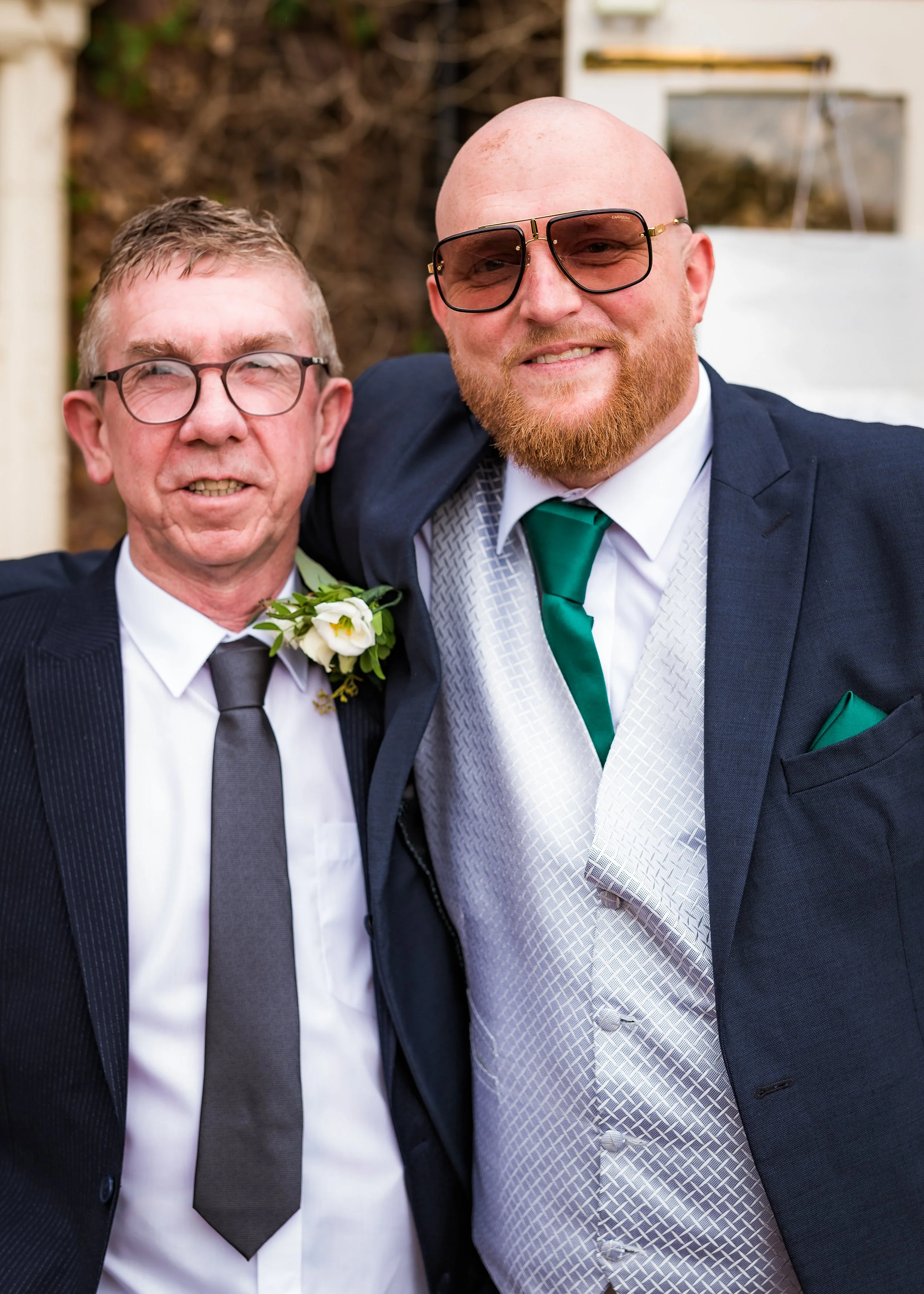 Two men in formal suits and glasses, one with a beard and the other clean-shaven, smiling at a social event.