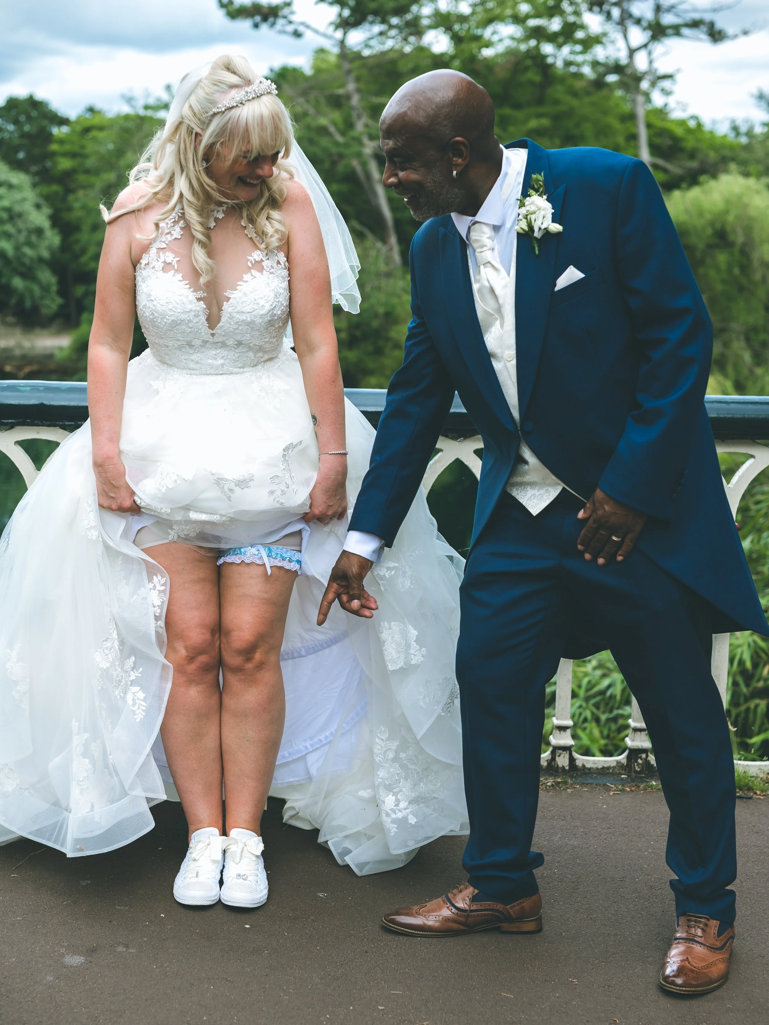 A bride in a wedding dress and a groom in a suit are holding hands and laughing, standing outdoors with greenery in the background.