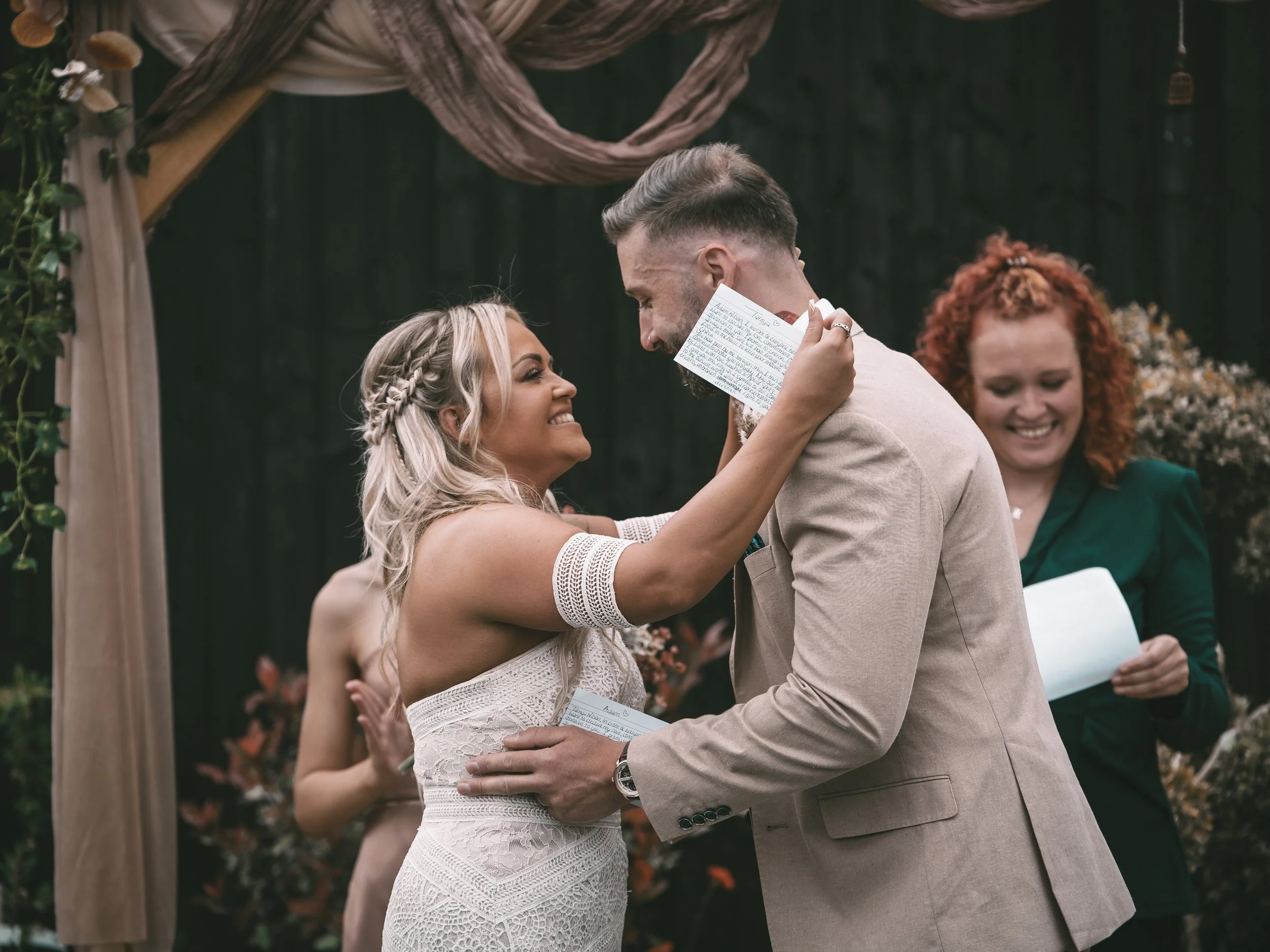 A couple exchanging vows at a wedding ceremony outdoors, with a woman holding a handwritten note and a woman in a green dress standing in the background.