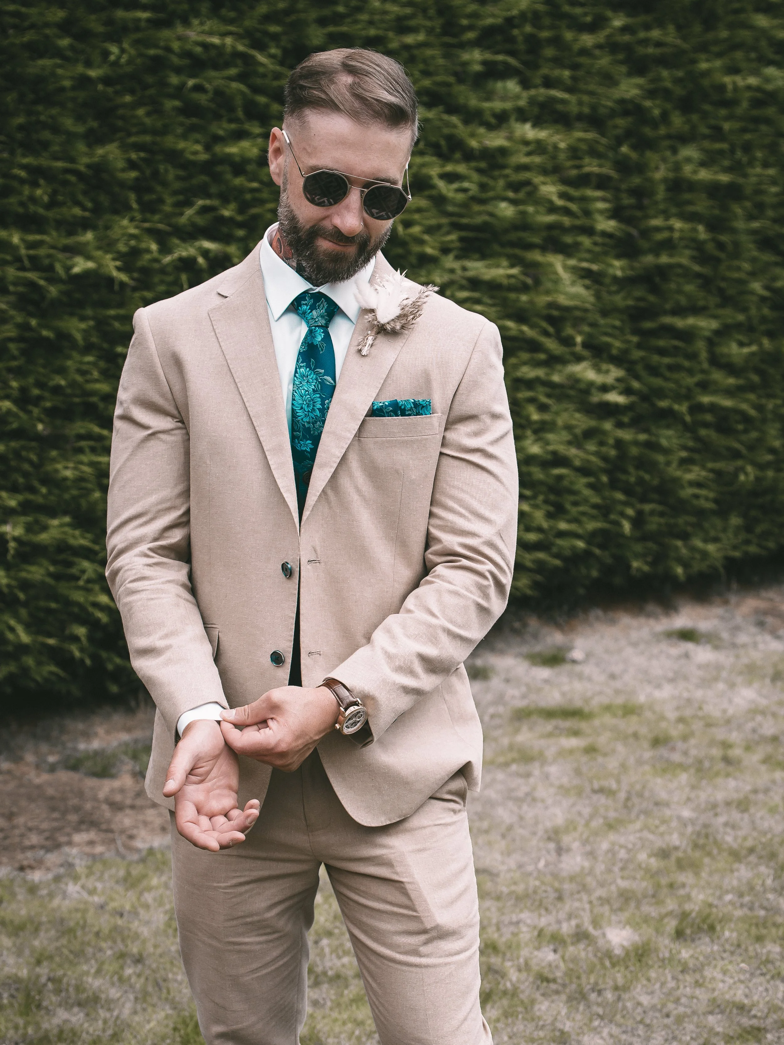 A man in a beige suit with a blue patterned tie, wearing sunglasses, stands outdoors in front of green bushes. He has a feather boutonniere on his jacket's lapel and is holding hands with someone not fully visible in the image.