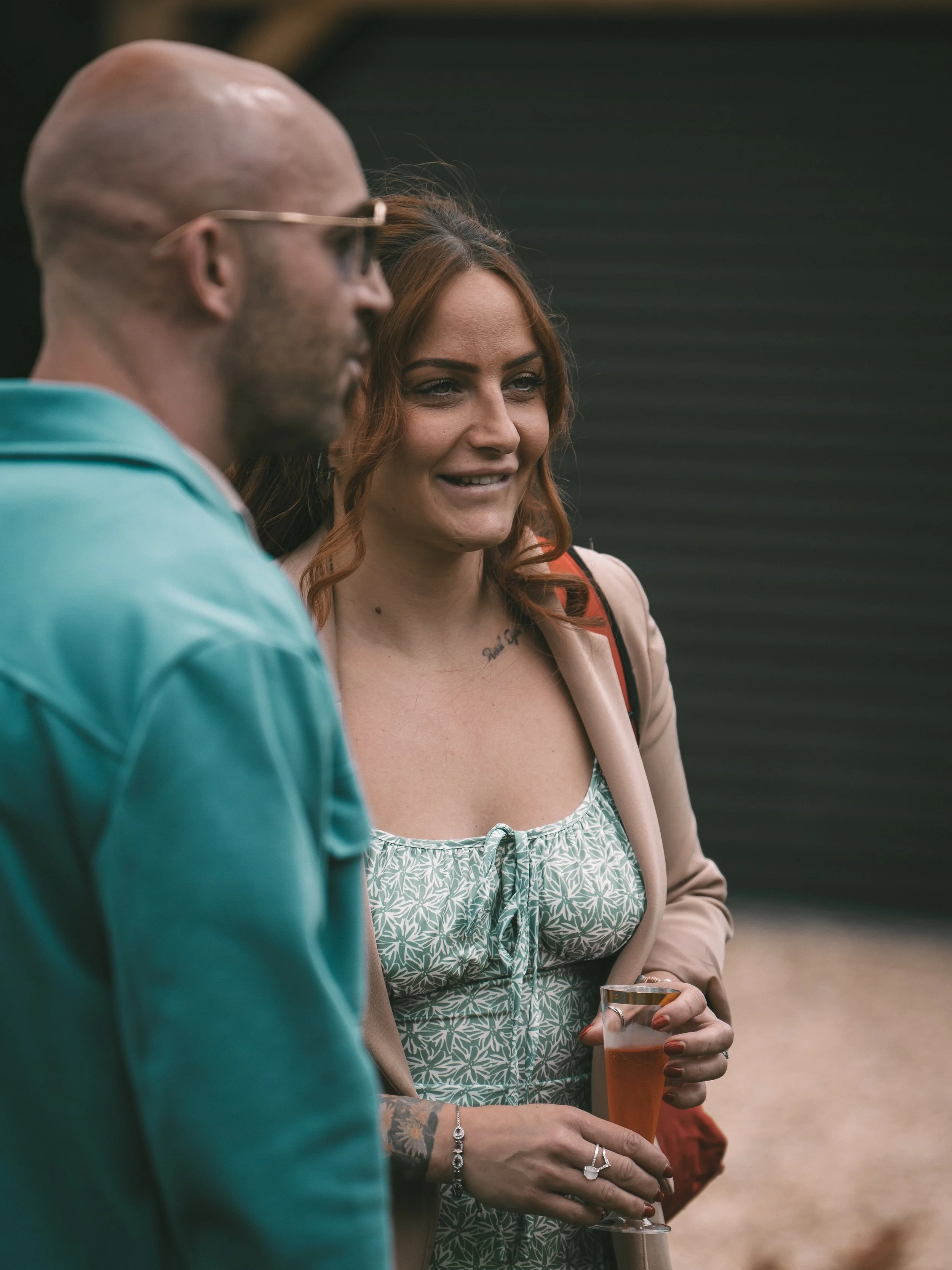 A woman with red hair, wearing a beige blazer and a green patterned dress, holding a glass of beer while talking with a bald man in a teal jacket, outdoors.