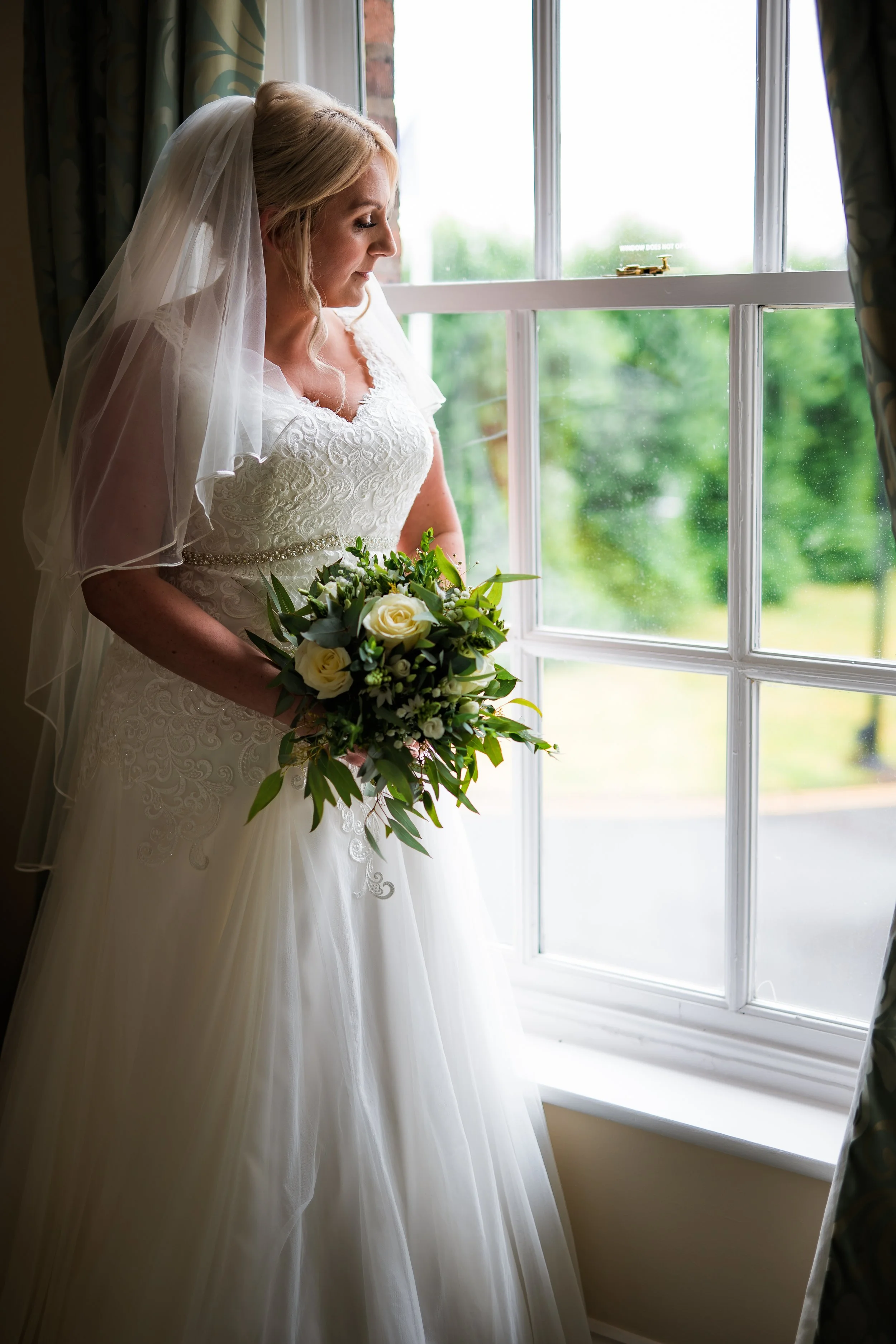 A bride with blonde hair, wearing a white lace wedding dress and veil, holding a bouquet of white roses and greenery, standing near a window with greenery outside.