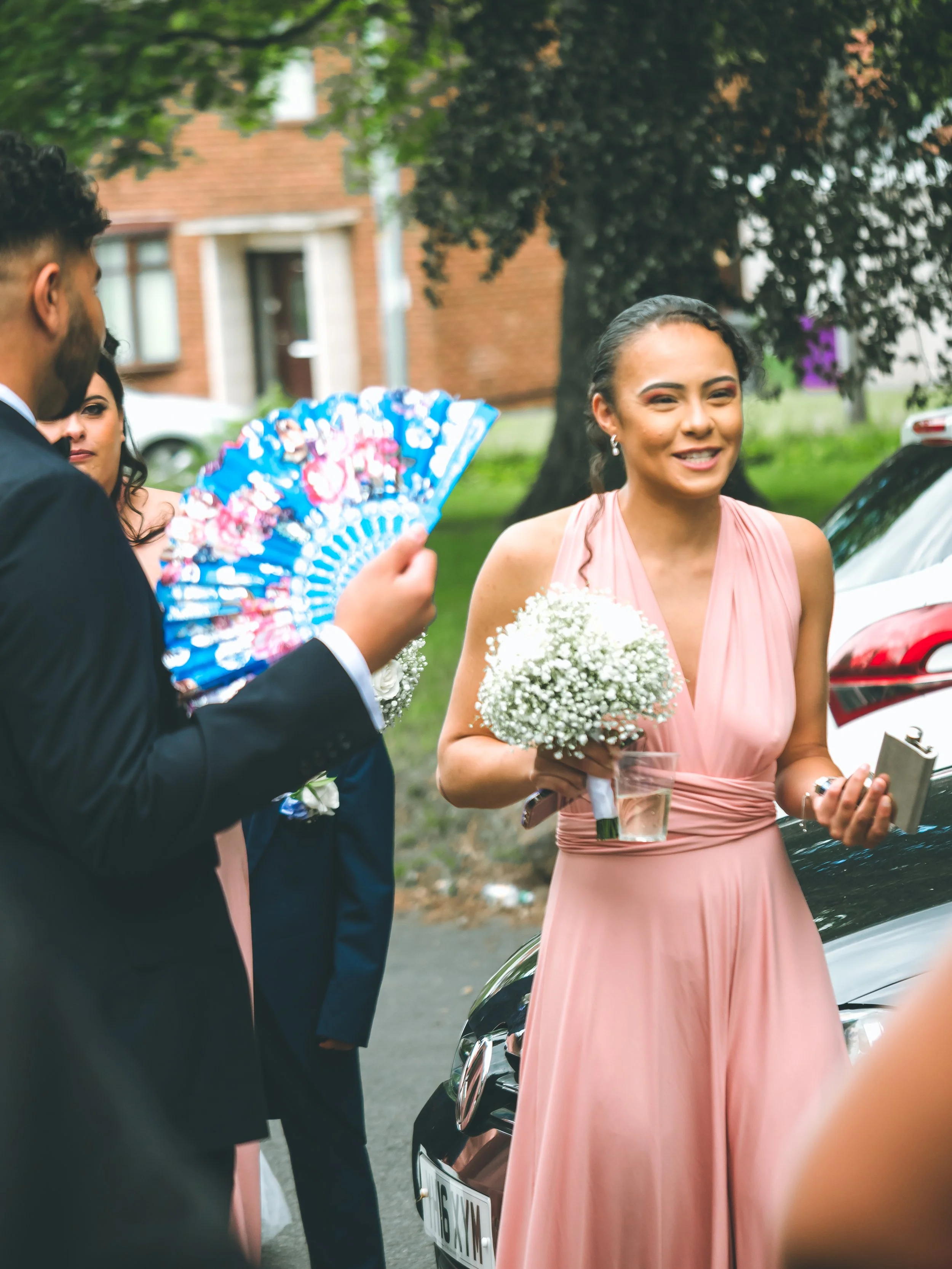 A woman in a pink dress smiling and holding a bouquet of white flowers, standing next to a man in a suit, with a woman holding a colorful fan nearby, outdoors near a parked car and trees.