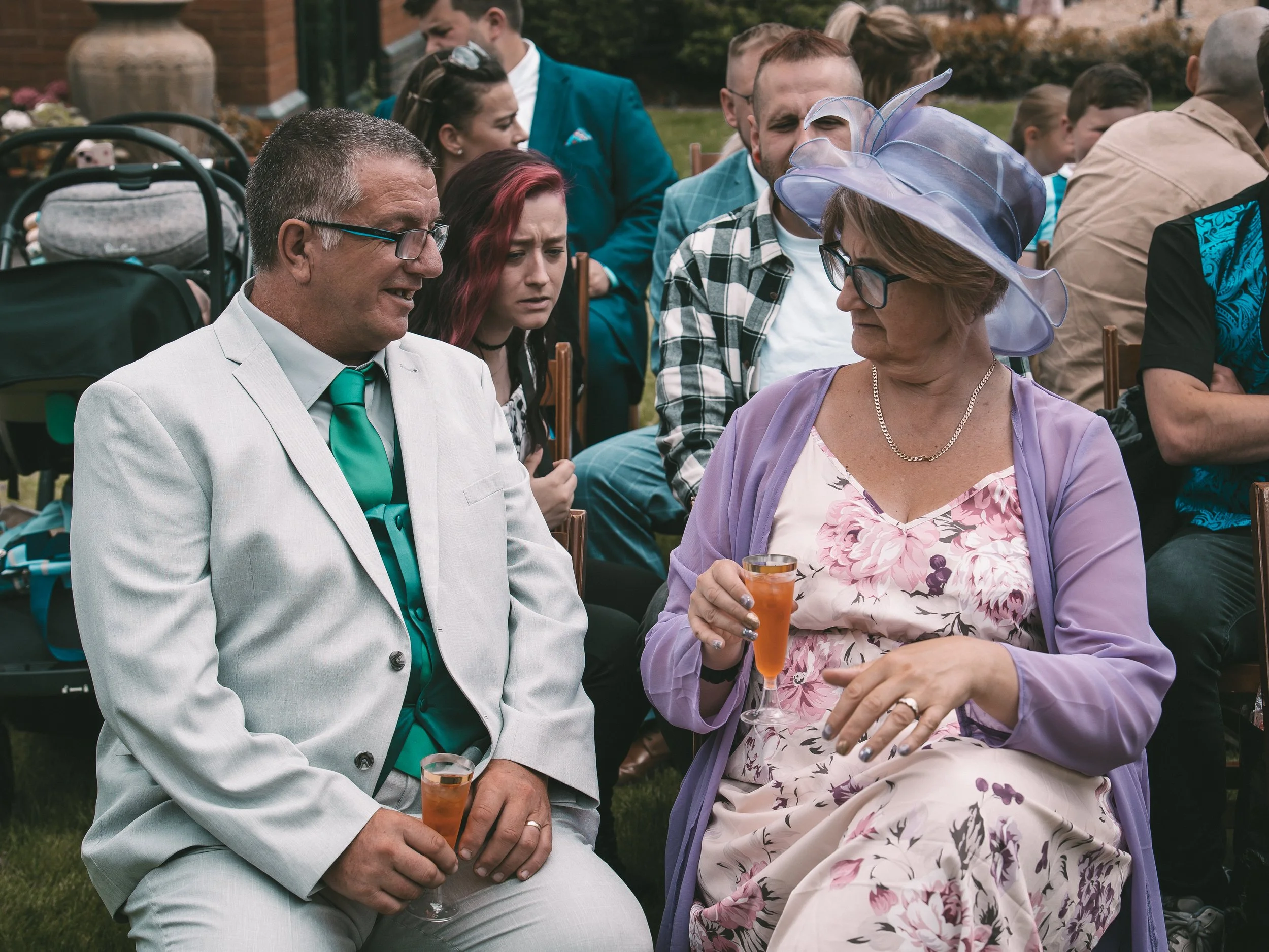 A group of people attending an outdoor event, seated on chairs, socializing. Prominent in the image are an older man wearing a light suit with a green tie and glasses and an older woman wearing a purple hat, glasses, and a floral dress holding a drin