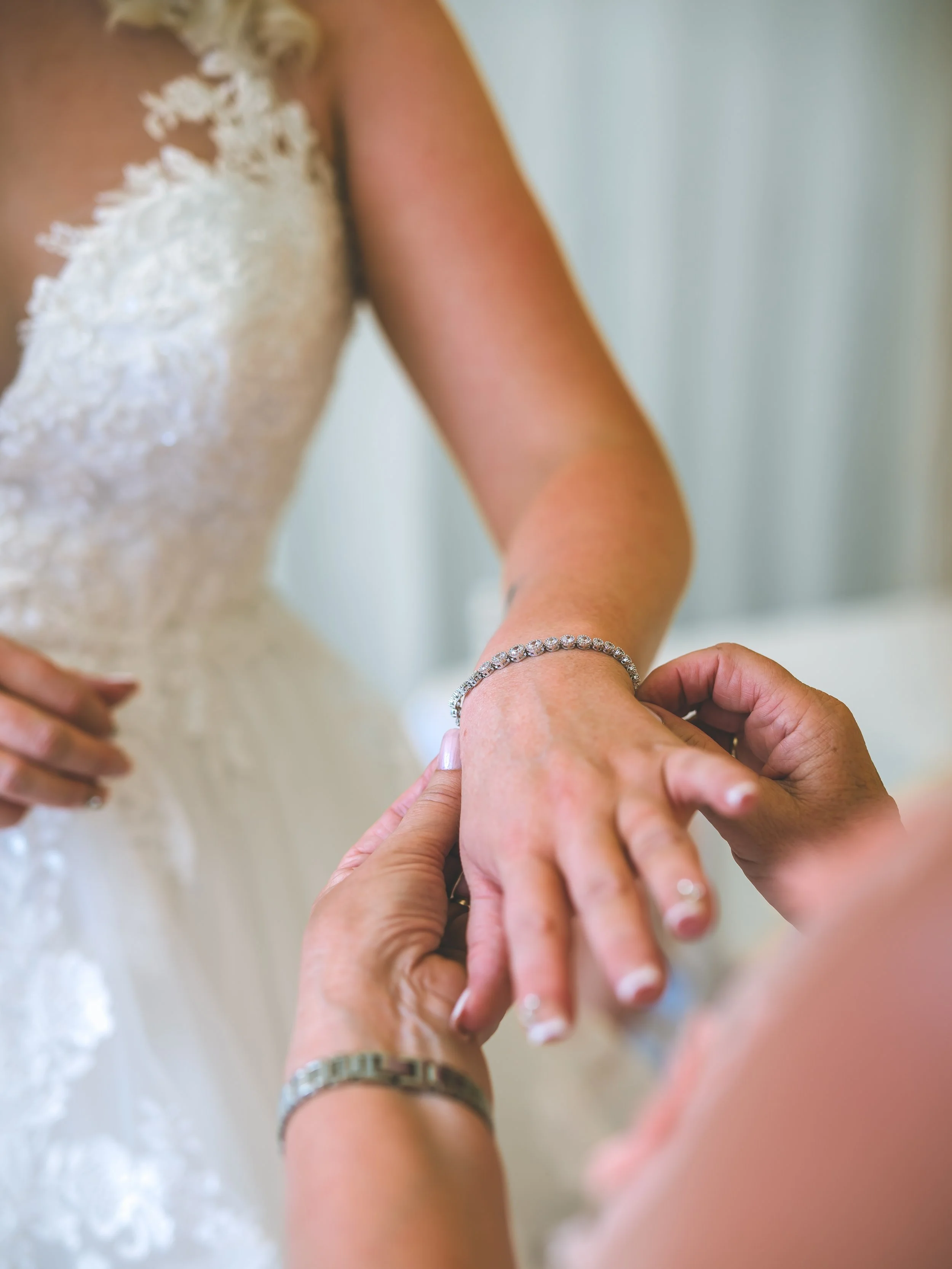 A woman in a wedding dress is putting a ring on her finger as another person helps her. The focus is on their hands and jewelry.