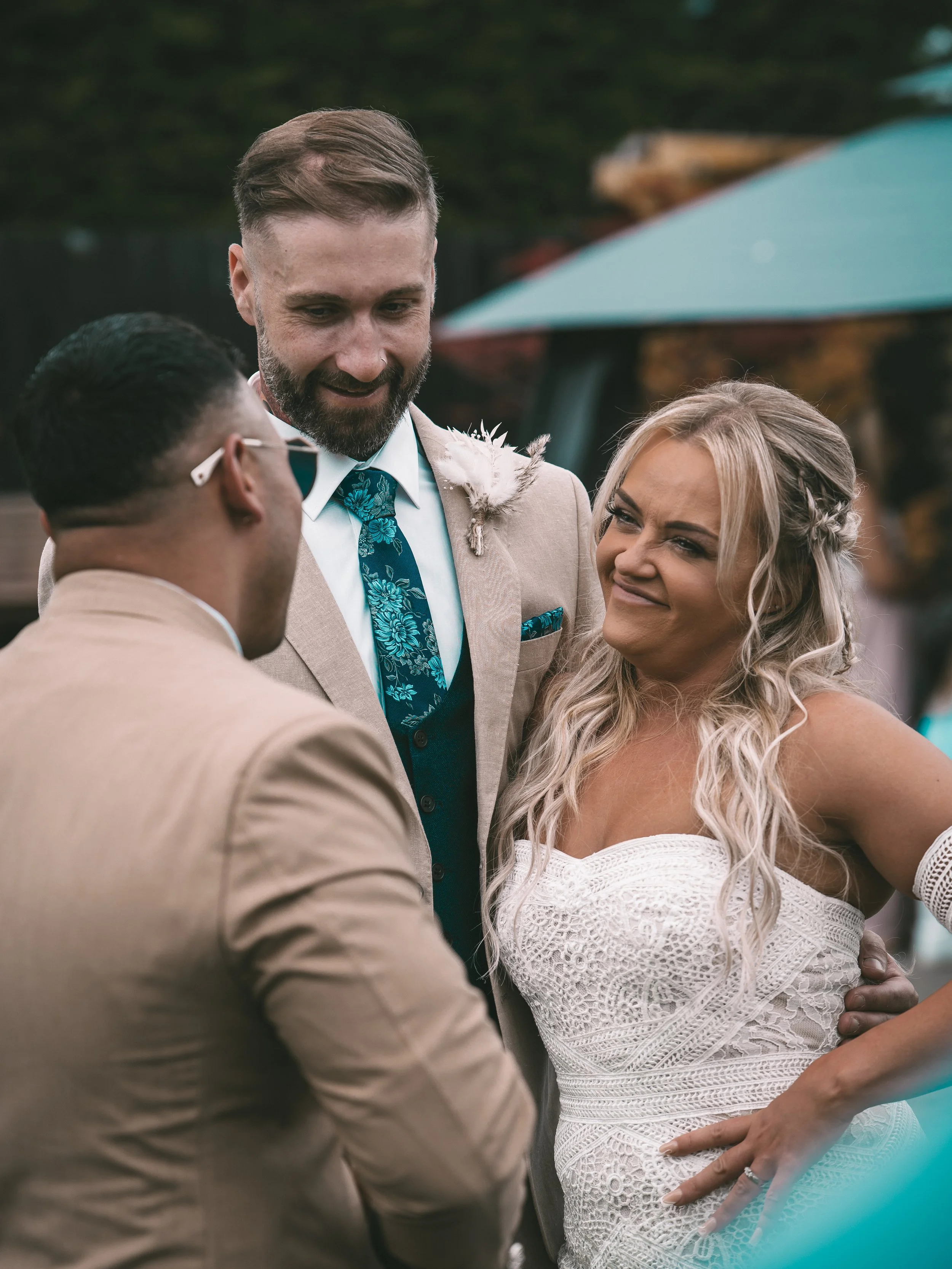 A wedding ceremony outdoors, with a bride and groom holding hands and looking at each other, the groom wearing a beige suit and the bride in a white lace dress, with a man in glasses and a beige jacket officiating the ceremony.