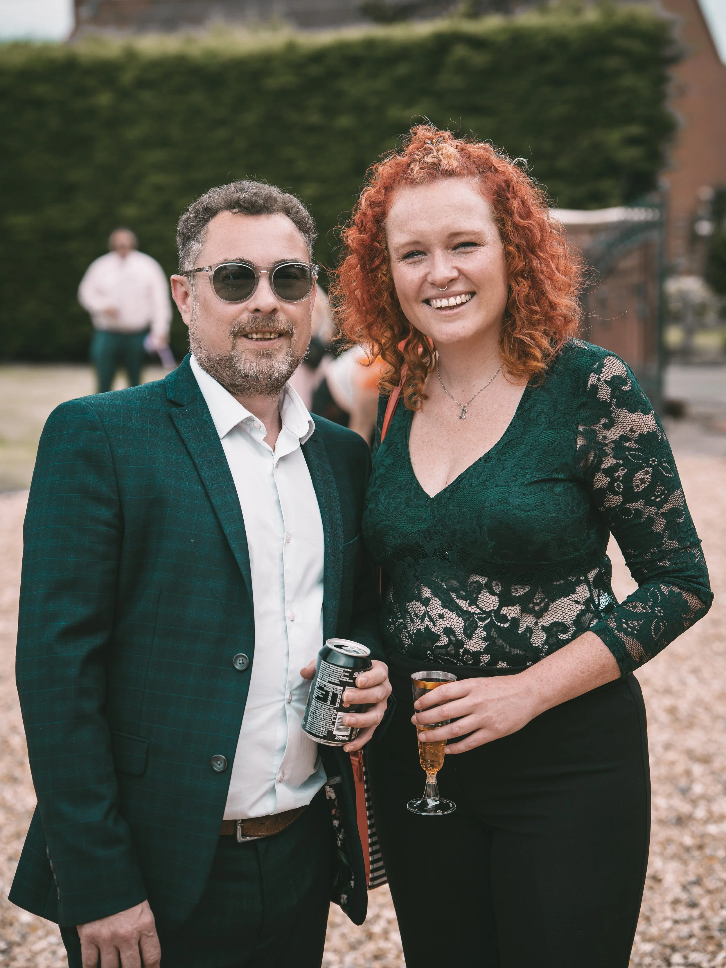 A man and woman smiling at an outdoor gathering, holding drinks. The man wears sunglasses, a dark blazer, and a white shirt. The woman has curly red hair, a lace green top, and a nose ring.