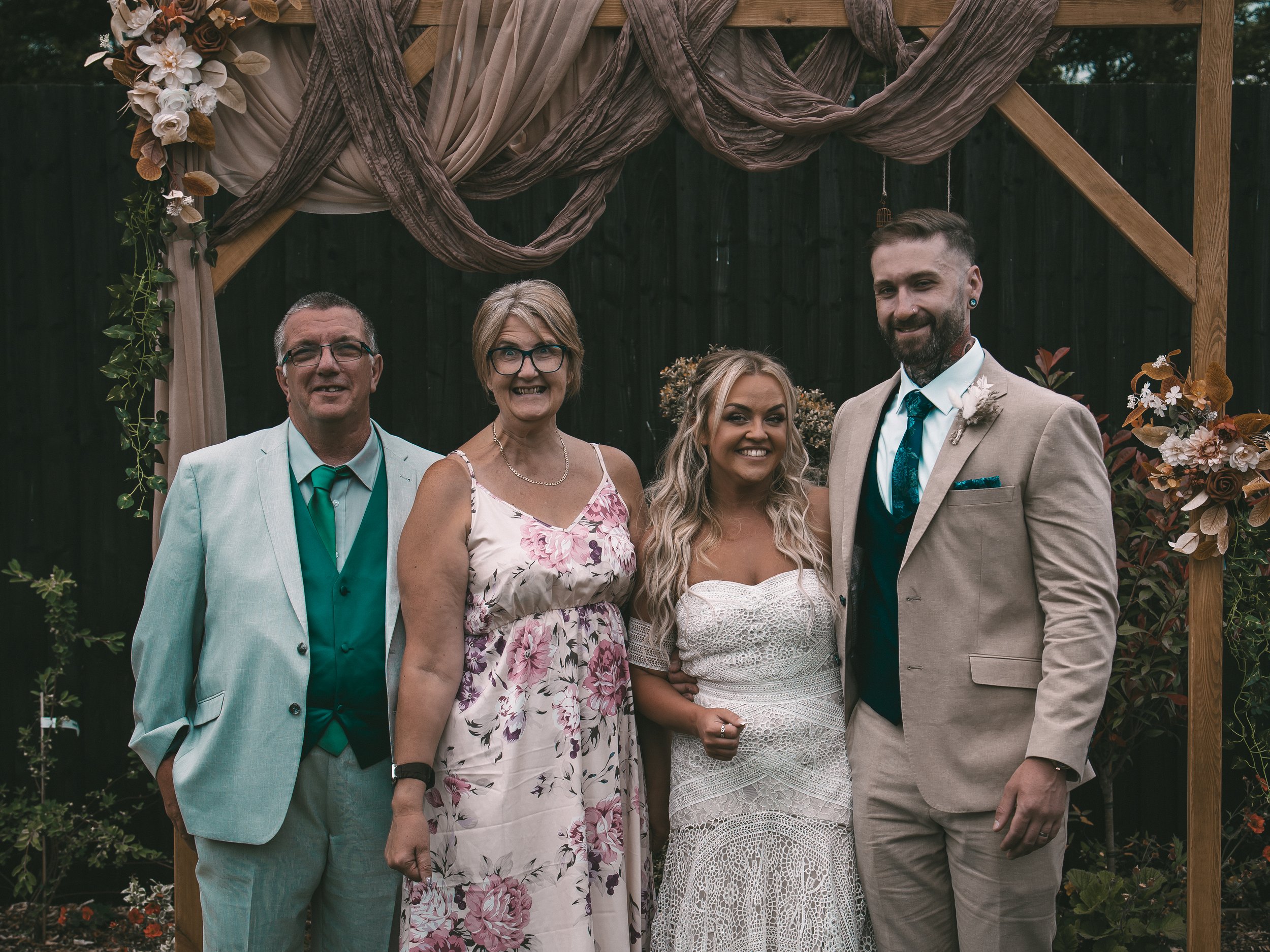 Group of five people at a wedding, standing under a decorated wooden arch with draped fabric and flowers, smiling at camera.